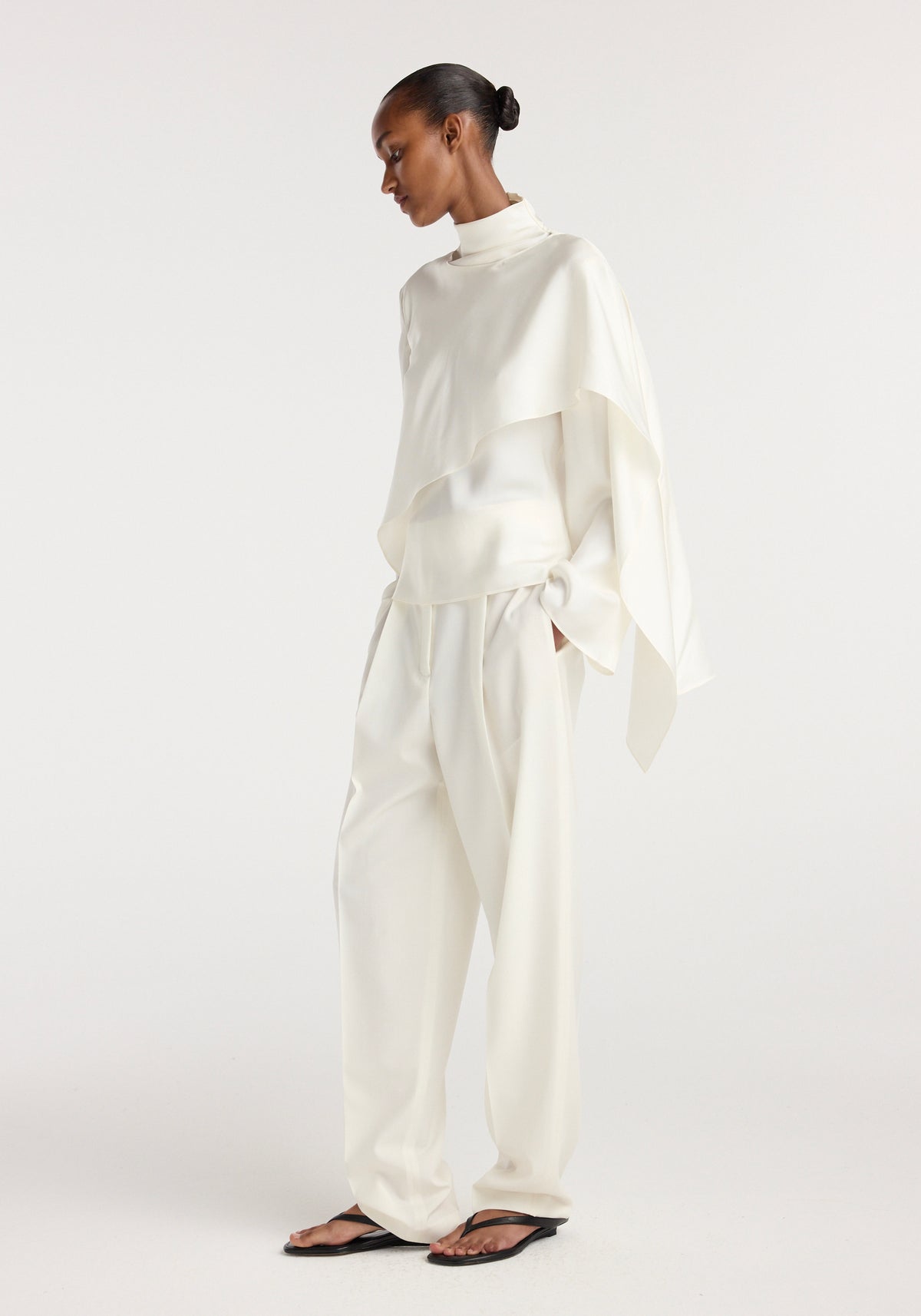 Model wearing a cream silk foulard top with a high neck and asymmetrical draped design paired with matching wide-leg trousers and black sandals, standing against a plain background.