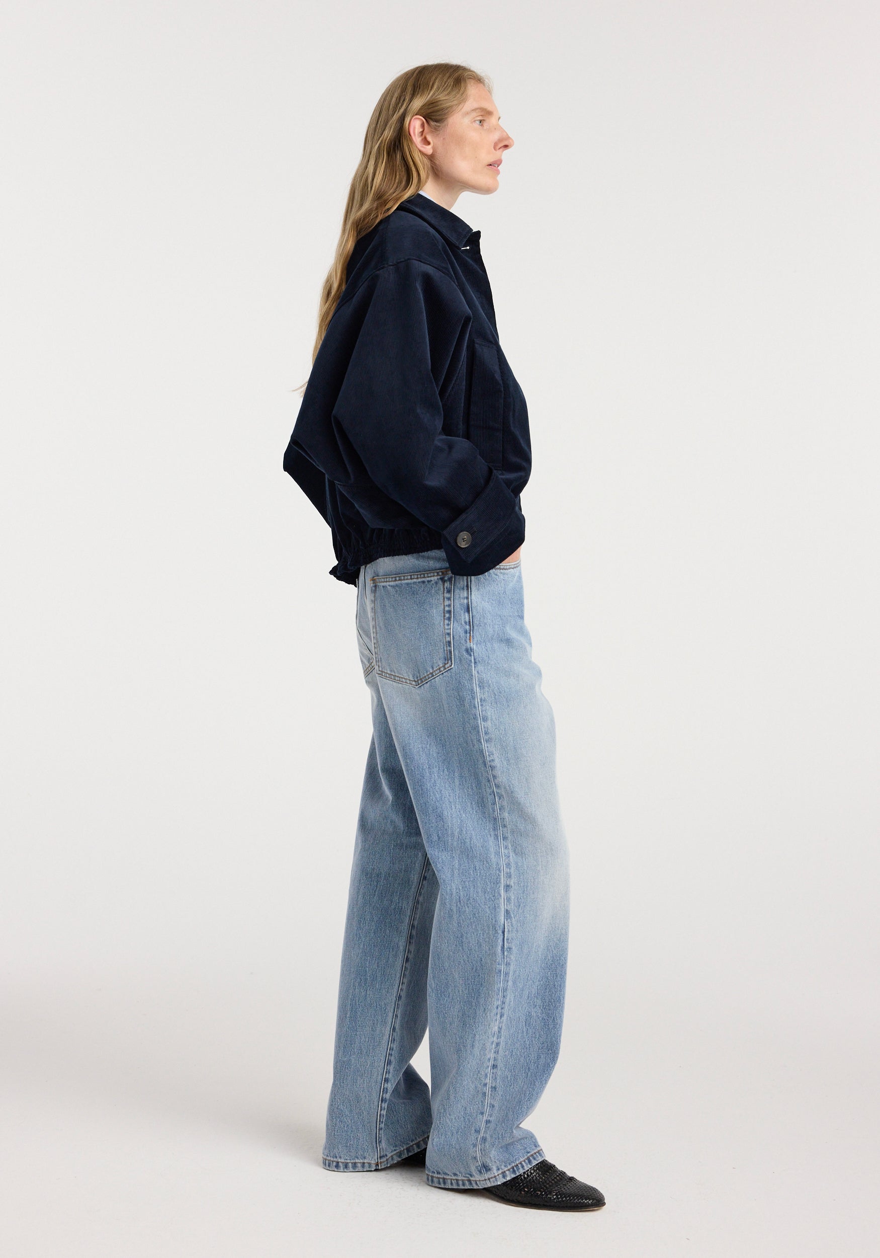 Side view of a woman wearing a dark navy Corduroy blouson jacket | night paired with light blue wide-leg jeans and black shoes against a plain background.