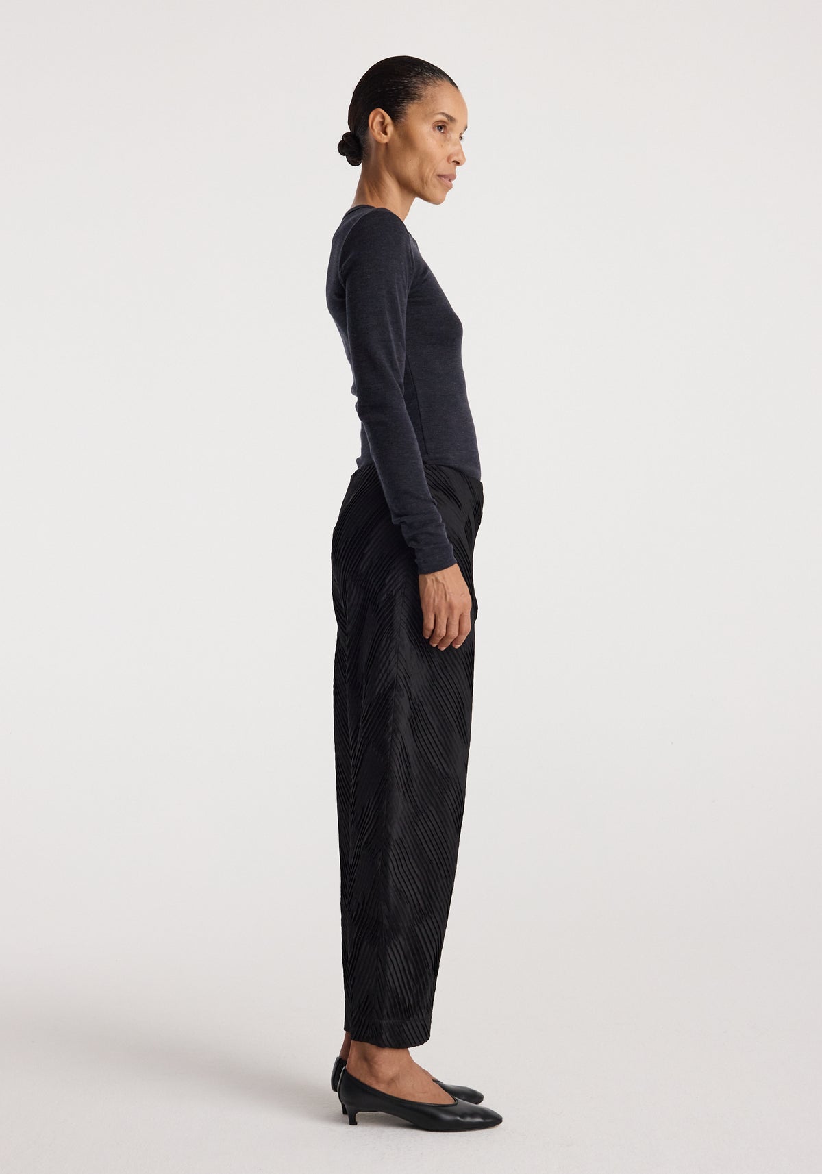 Side view of a woman wearing a fitted fine merino long sleeve | anthracite melange top paired with black pants and black low-heeled shoes against a plain background.
