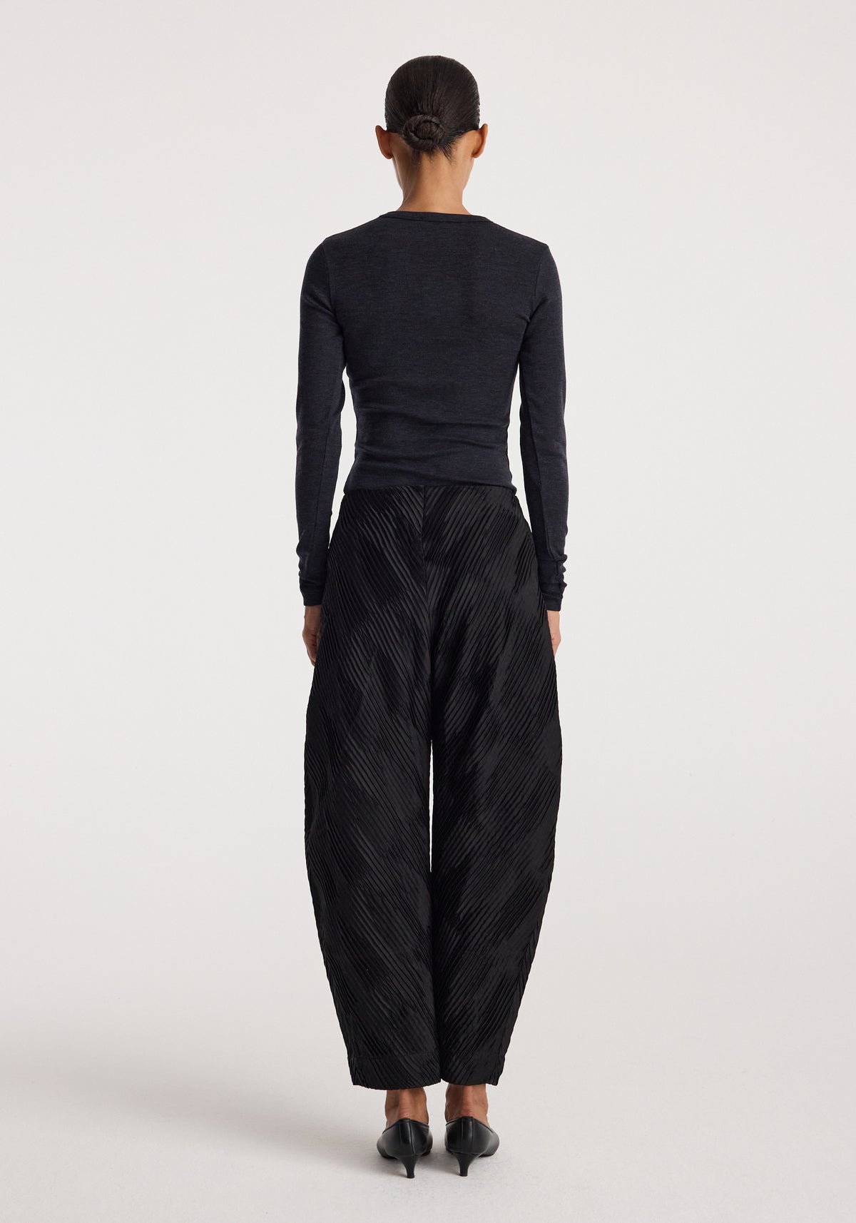 Woman seen from the back wearing a fitted dark grey long sleeve top and loose black textured wide-leg pants with black heeled shoes, featuring the Fine merino long sleeve | anthracite melange.