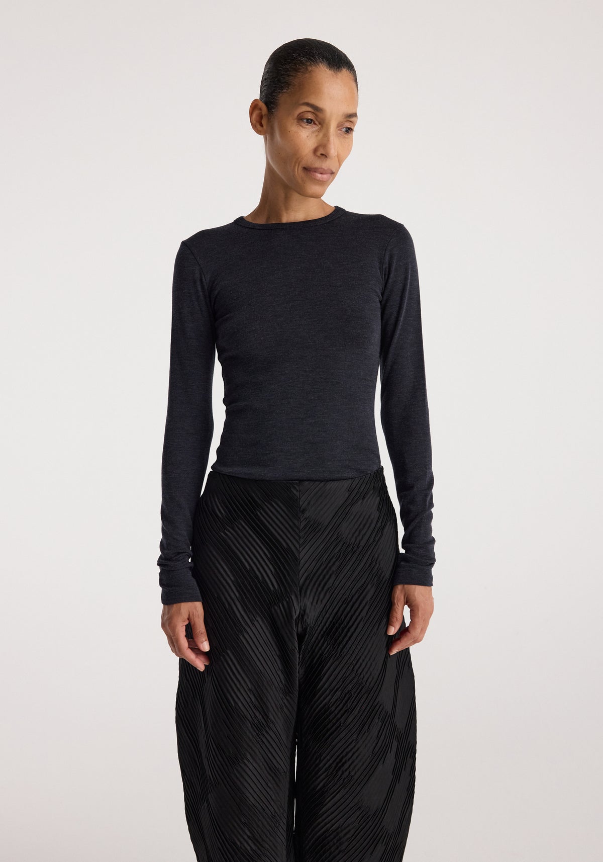 Woman wearing a fitted Fine merino long sleeve | anthracite melange top with long sleeves, paired with loose black textured pants, standing against a plain background.