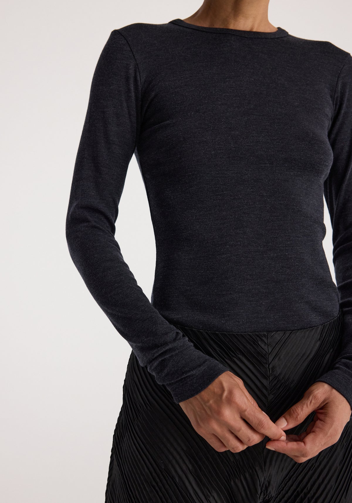 Close-up of a person wearing a fitted Fine merino long sleeve | anthracite melange sweater with a round neckline and long sleeves, paired with a black pleated skirt.