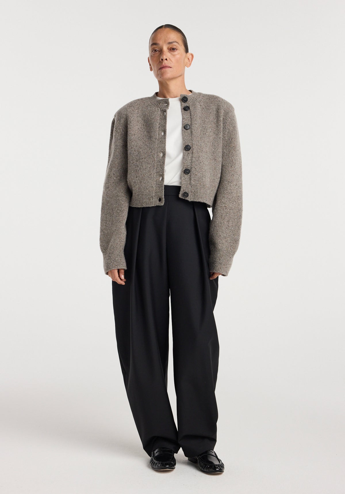 Woman wearing a buttoned Statement mouliné cardigan | dark taupe melange with long sleeves over a white top, paired with loose black trousers and black shoes, standing against a plain background.
