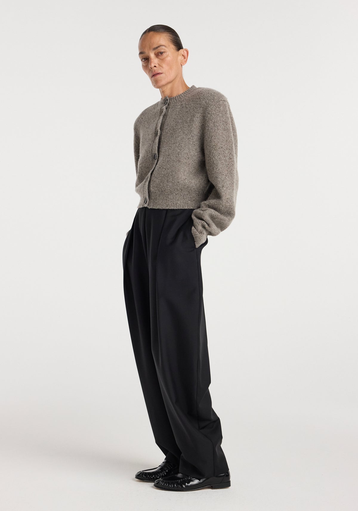 A woman wearing a Statement mouliné cardigan | dark taupe melange, styled with high-waisted black wide-leg pants and black shoes, standing against a plain background.