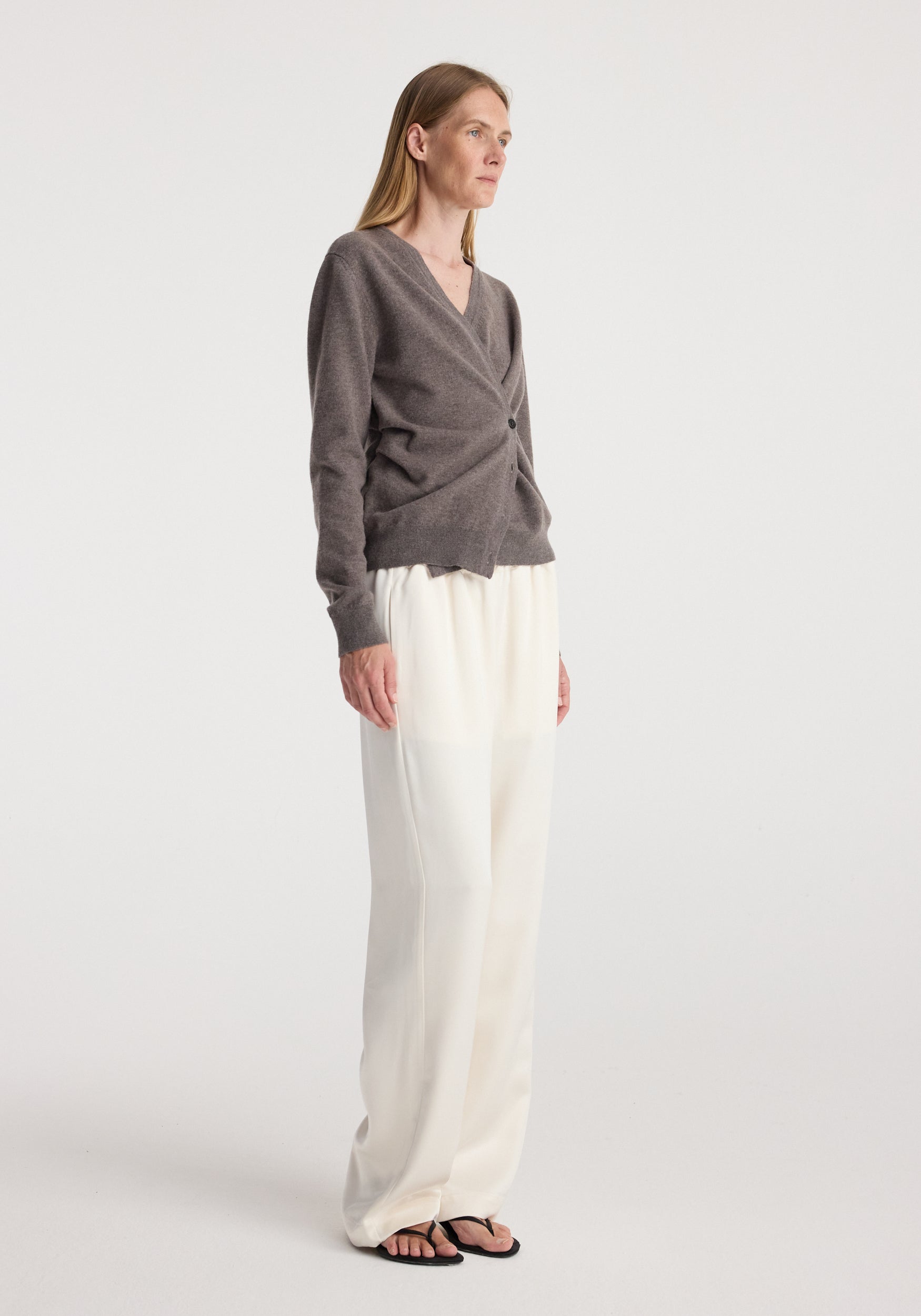 Woman wearing a mud-colored twisted ballet cardigan | mud with long sleeves and a V-neck, paired with loose white pants and black sandals, standing against a white background.