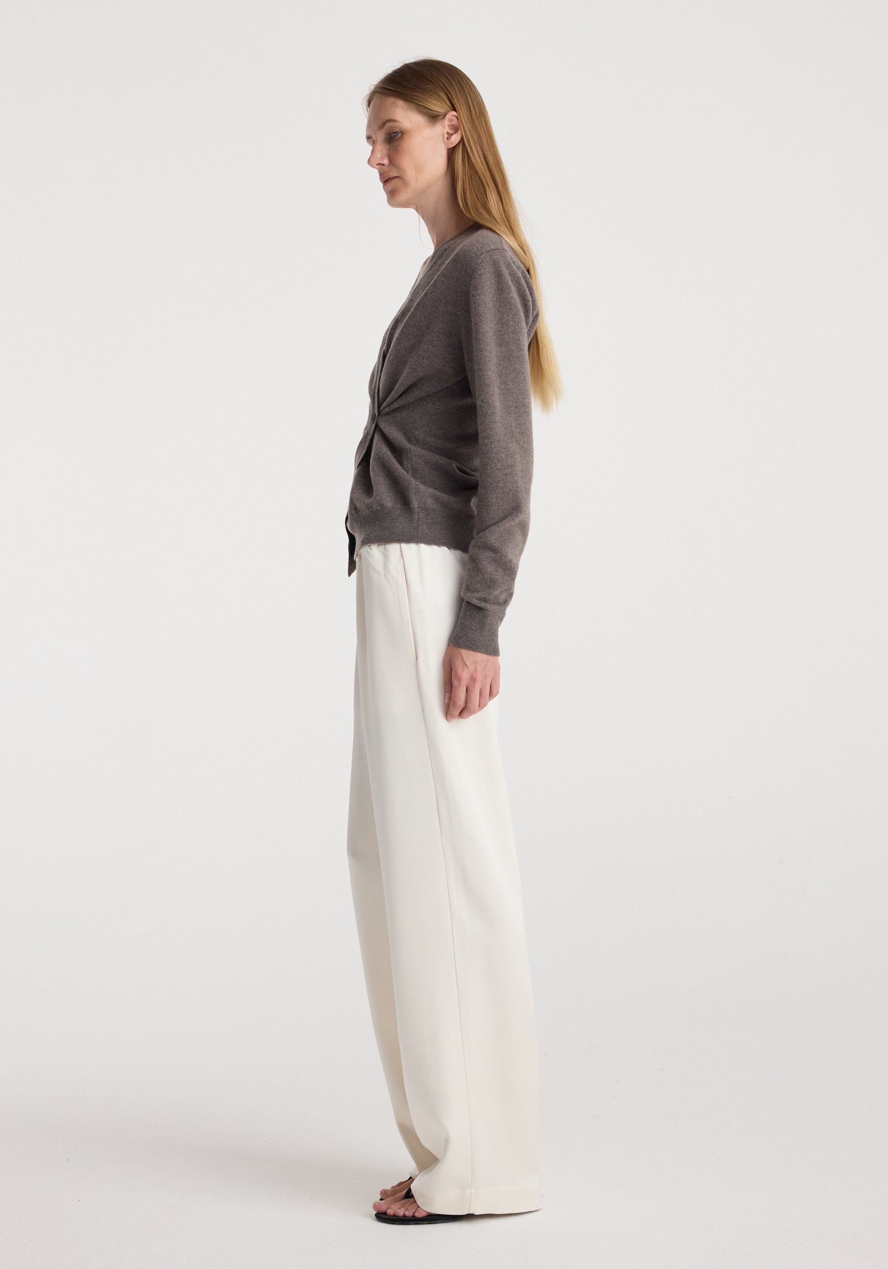 Woman wearing a twisted ballet cardigan | mud color with long sleeves, paired with wide-leg white pants and black open-toe sandals, standing in profile against a plain background.