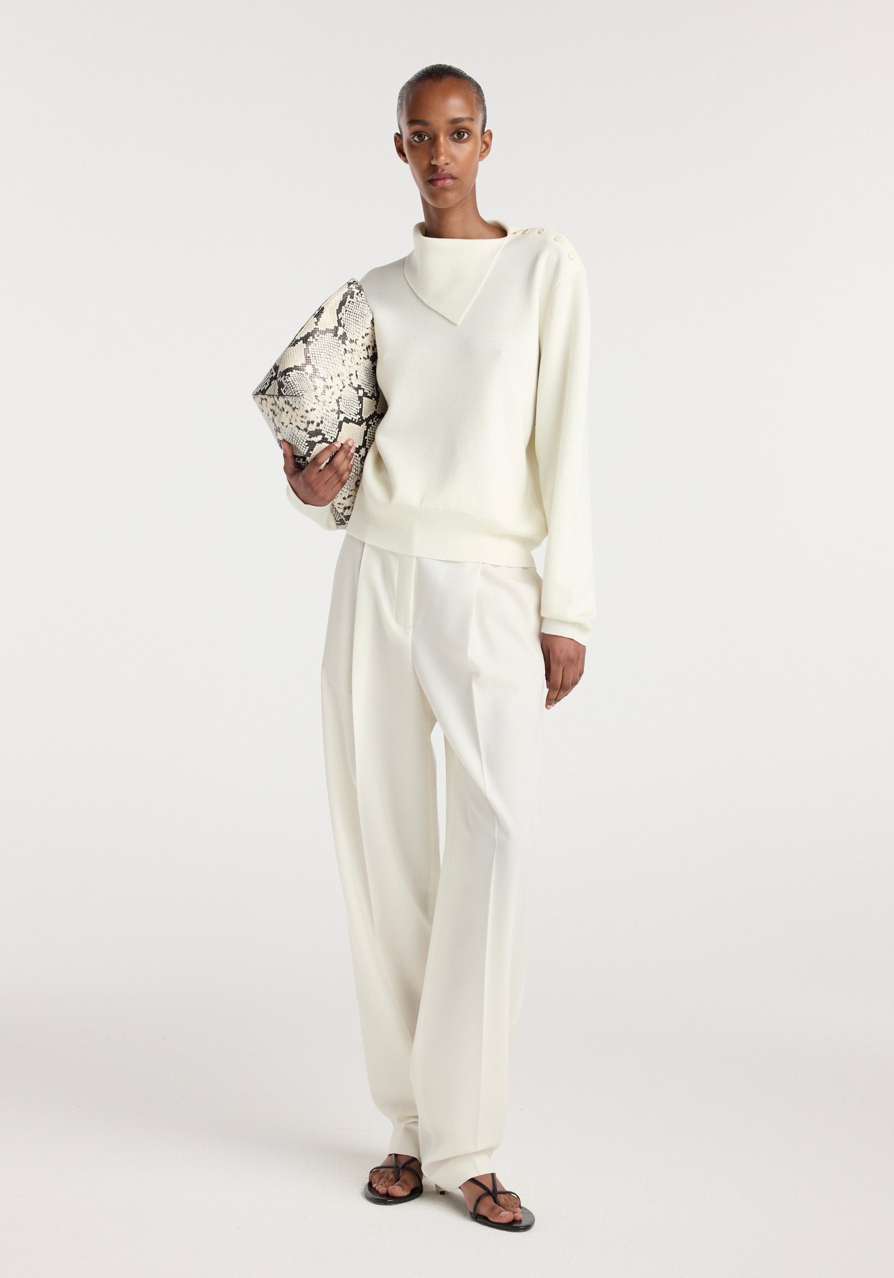 Model wearing an off white uneven collar polo paired with matching wide-leg trousers, carrying a snakeskin-patterned clutch against a plain background.