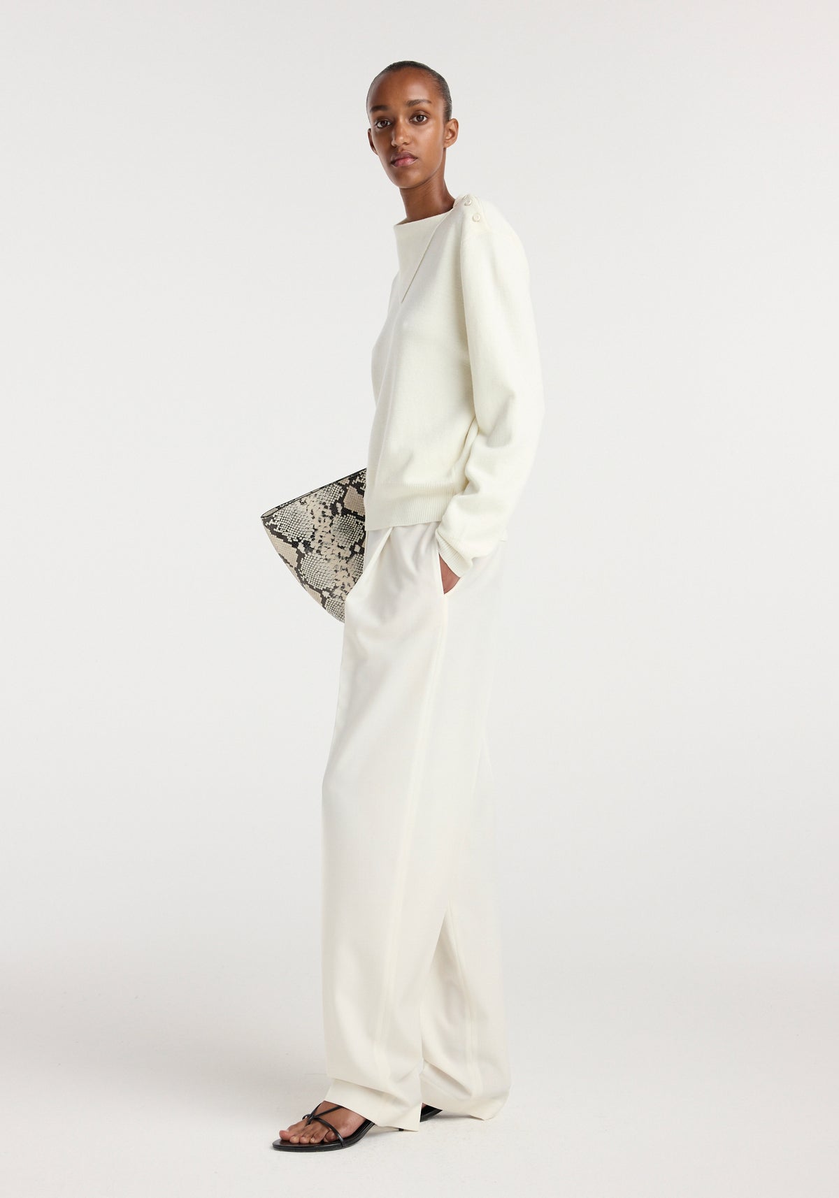 Model wearing an off white uneven collar polo paired with loose-fitting off white trousers, black strappy sandals, and holding a black and white snakeskin-patterned clutch against a plain background.
