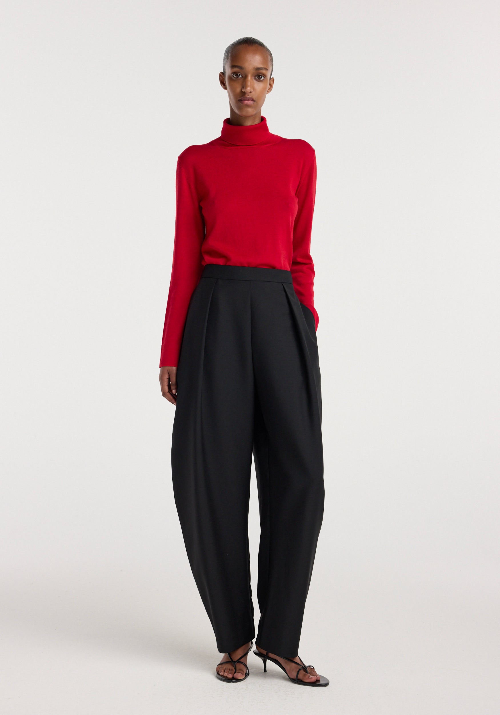 Woman wearing a red long-sleeve turtleneck top paired with black Puck - Sculptural tailored trousers | black and black open-toe heels against a plain white background.