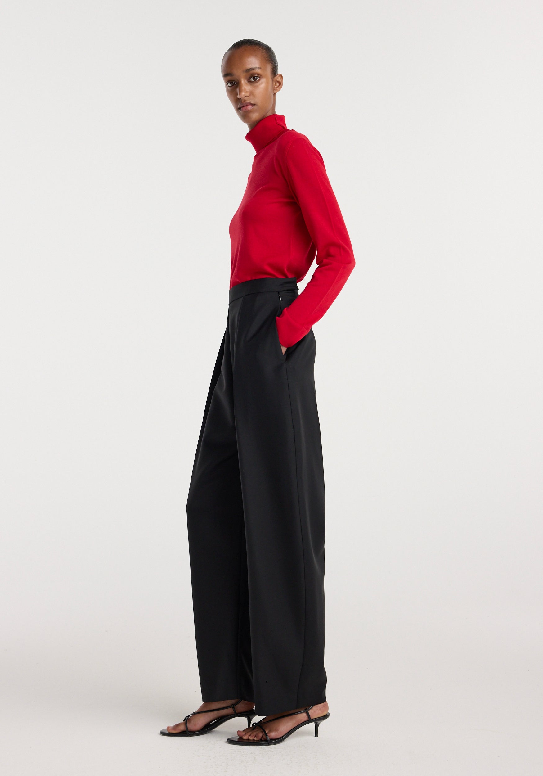 Model wearing black Puck - Sculptural tailored trousers | black styled with a red long-sleeve turtleneck top and black open-toe heeled sandals against a plain white background.