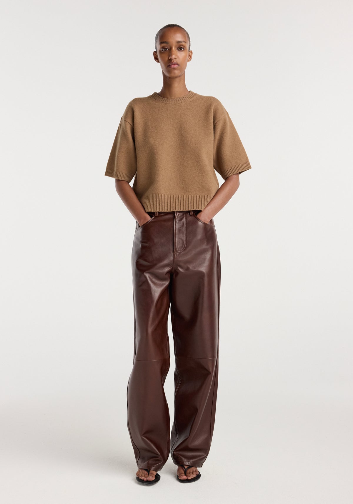 A model wearing Bruun - Signature baggy leather trousers | tobacco, paired with a brown short-sleeve knit sweater and black sandals, standing against a plain white background.