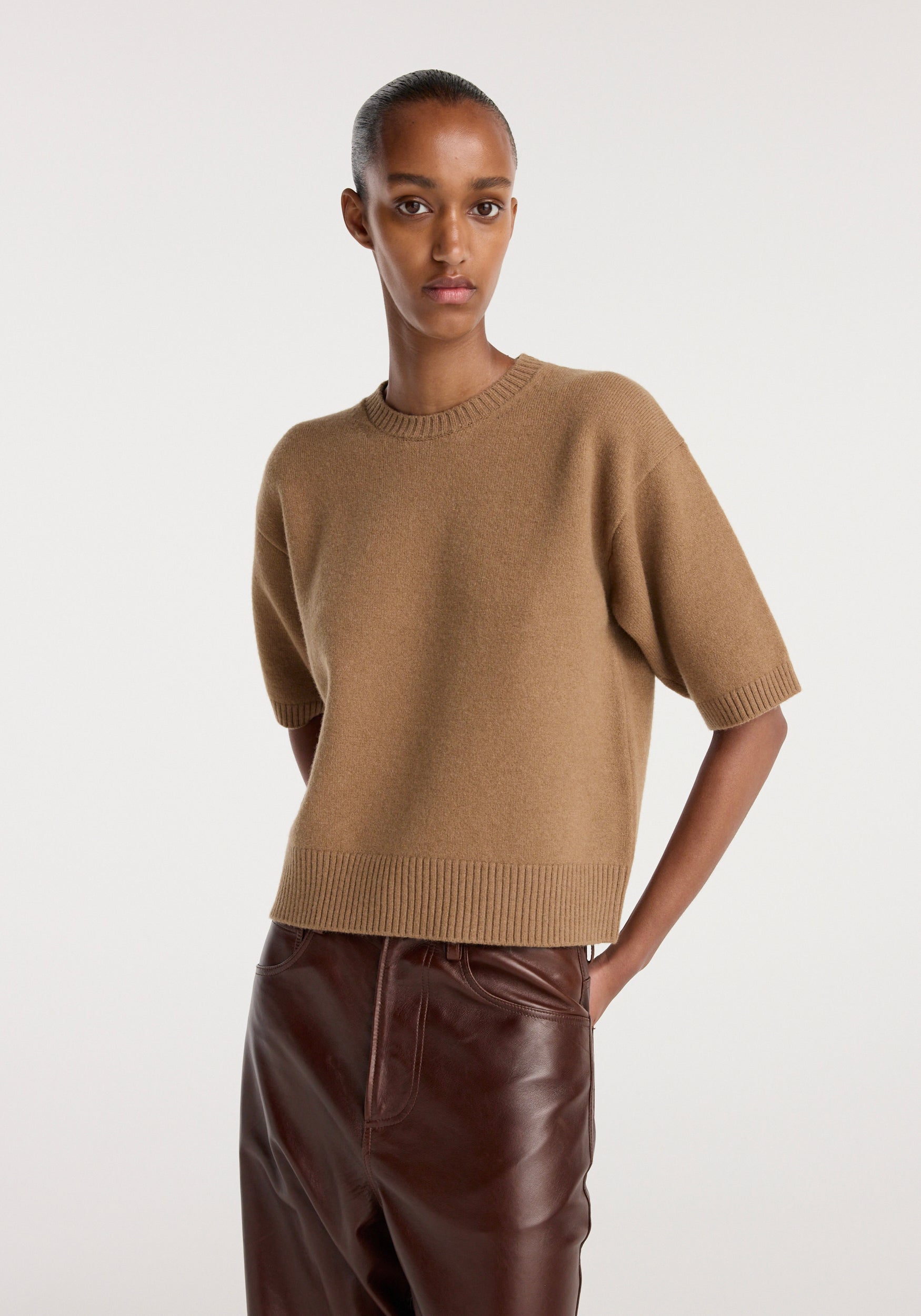 A woman wearing a camel-colored compact knitted top with short sleeves and ribbed detailing at the hem and cuffs, paired with dark brown leather pants against a neutral background.