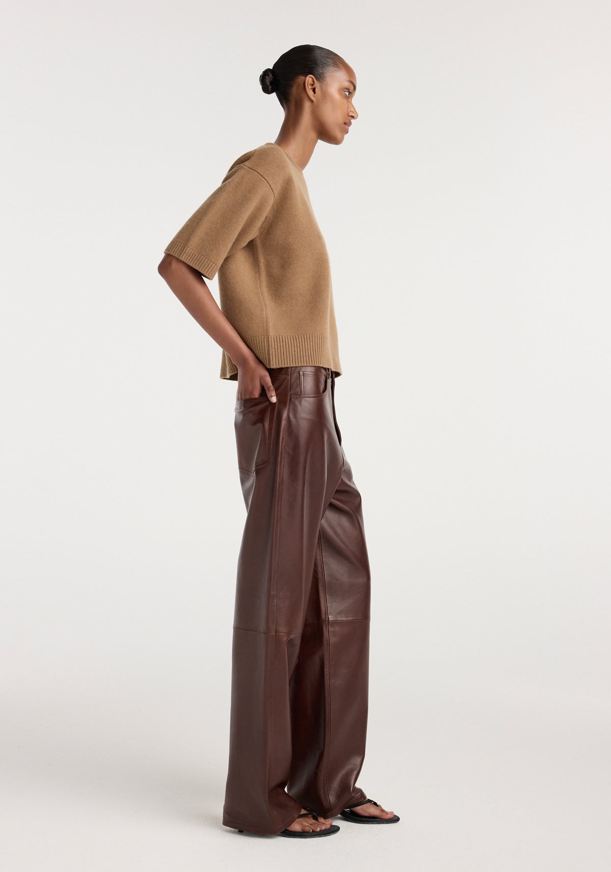 Side view of a woman wearing Bruun - Signature baggy leather trousers | tobacco paired with a loose tan short-sleeve sweater and black sandals against a plain background.
