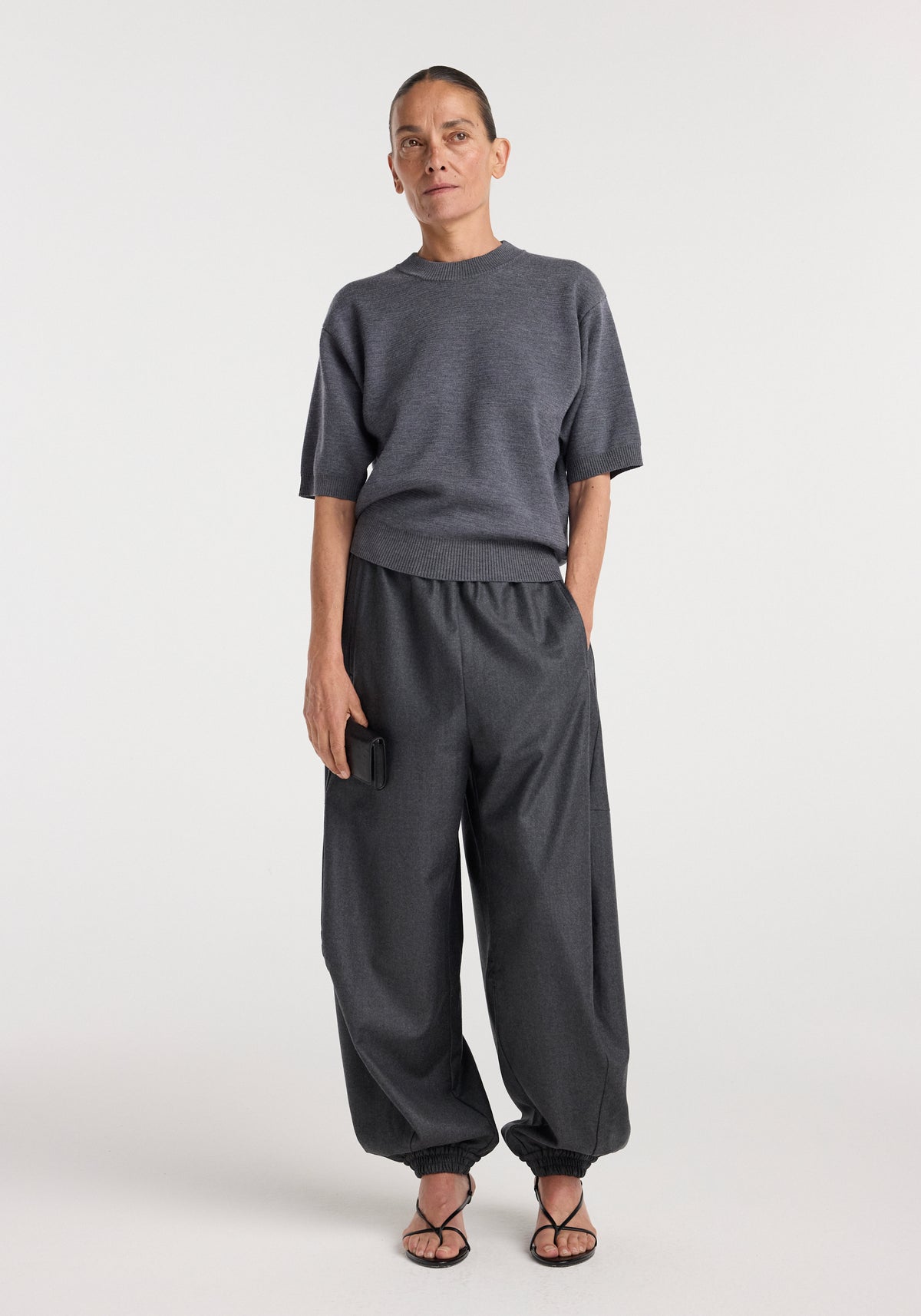 Woman wearing dark grey melange balloon track pants with elasticated cuffs, paired with a matching short-sleeve sweater and black open-toe sandals, standing against a plain background.