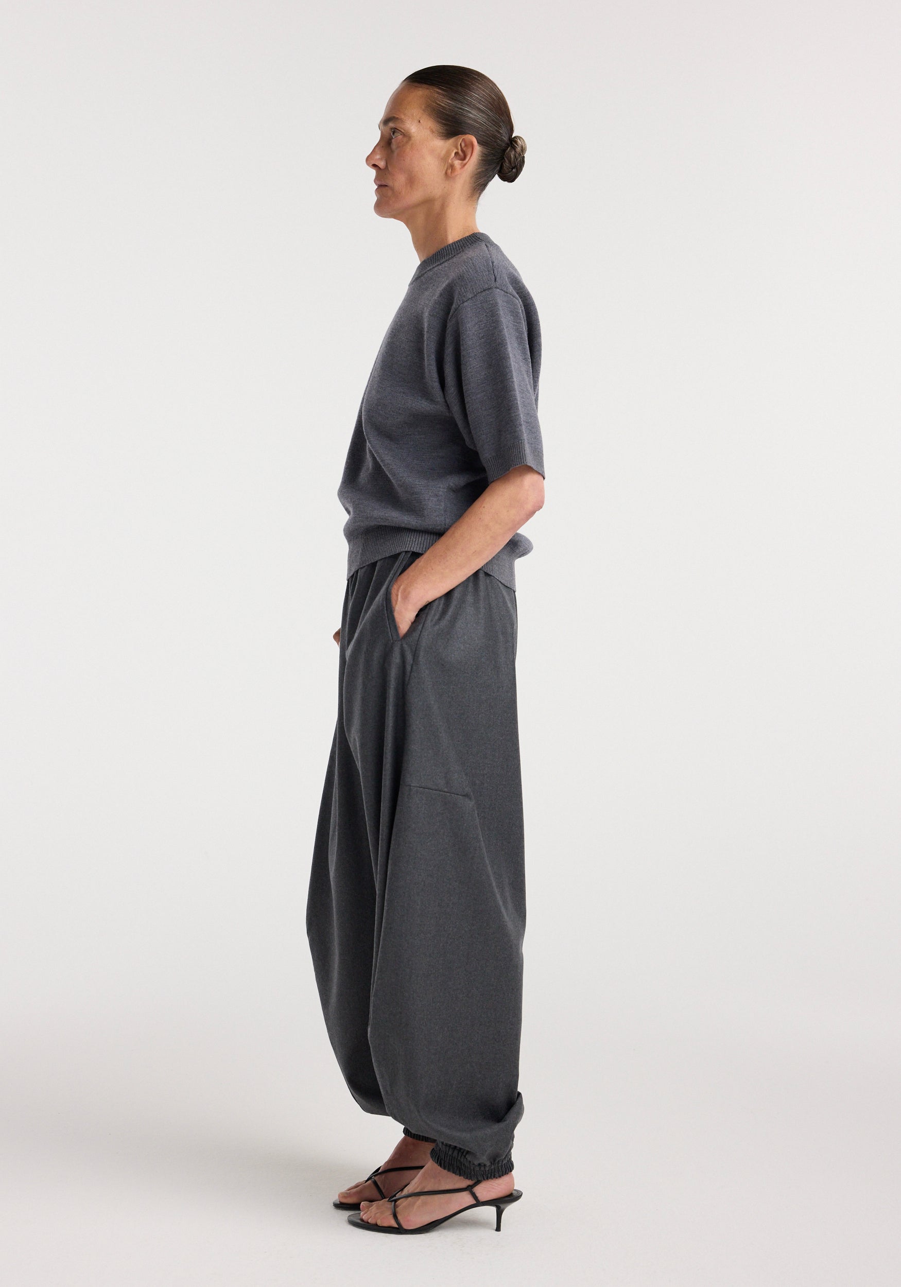 Side profile of a woman wearing Balloon track pants | dark grey melange, paired with a matching short-sleeve top and black strappy heels, standing against a plain background.