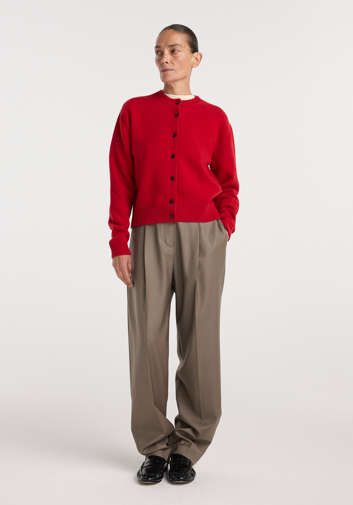 Woman wearing a Classic cardigan | red buttoned up, paired with loose beige trousers and black shoes, standing against a plain white background.