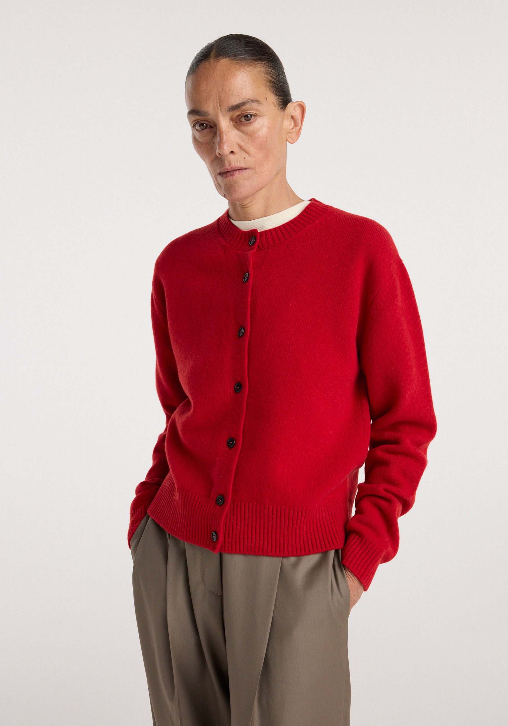 Woman wearing a bright red Classic cardigan | red with black buttons, paired with loose, beige trousers, standing against a plain white background.
