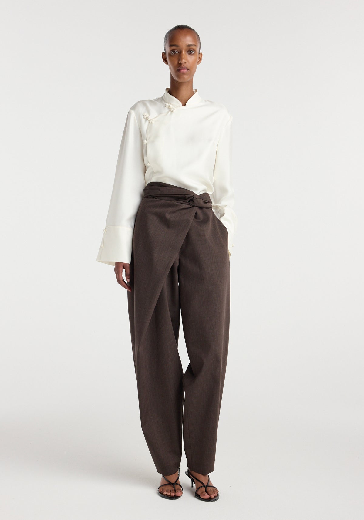 Model wearing Sarong knot trousers | dark brown, styled with a high waist, front overlapping knot detail, and straight leg fit paired with a white long-sleeve top and black strappy sandals.