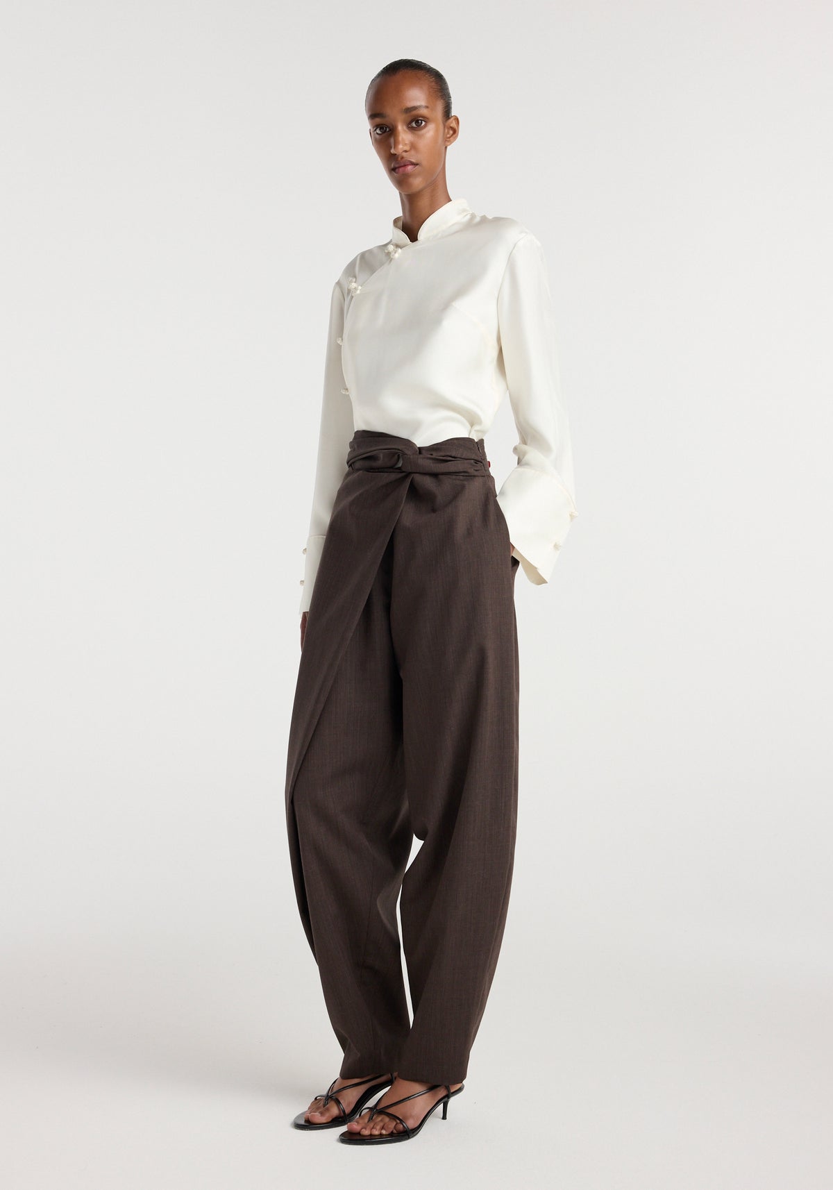 Model wearing Sarong knot trousers | dark brown paired with a white long-sleeve blouse and black open-toe heels against a plain background.