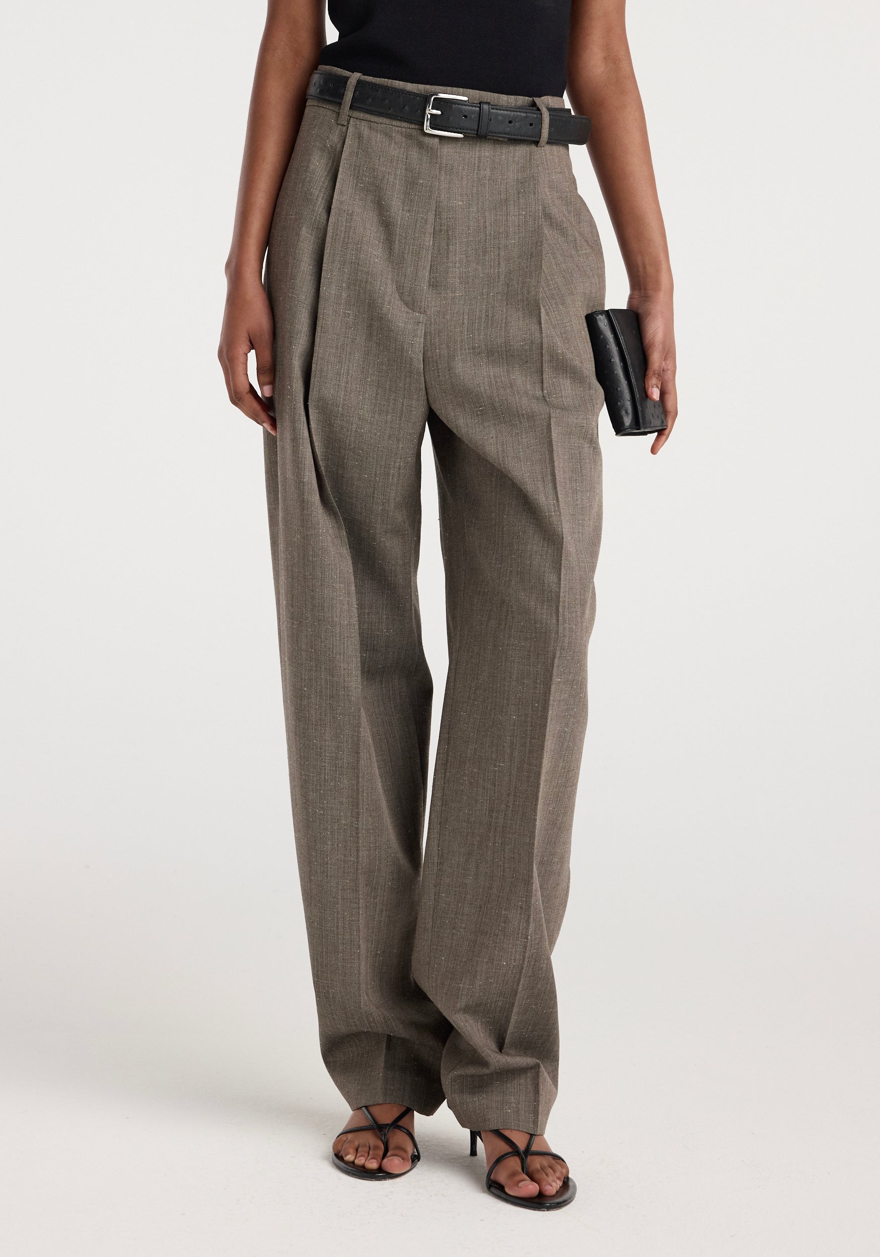 Front view of a model wearing Daria - Single pleat tapered trousers | earth slub in a muted earth tone, styled with a black belt, black top, black heeled sandals, and holding a black clutch purse.