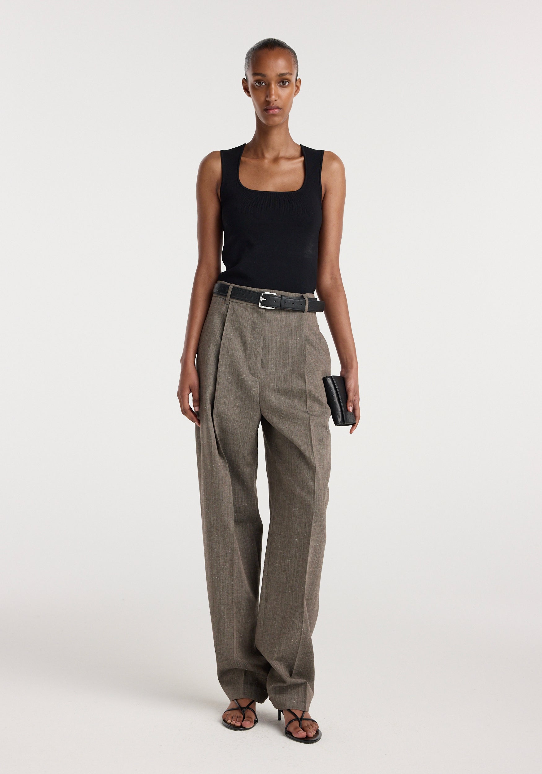 Woman wearing earth-tone textured single pleat tapered trousers with a black sleeveless top and black belt, styled with black sandals and a small clutch, showcasing Daria - Single pleat tapered trousers | earth slub.