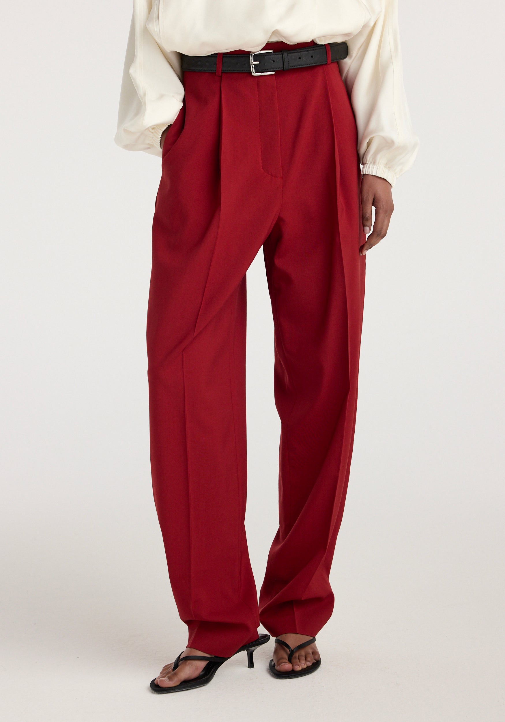 Front view of a model wearing Daria - Single pleat tapered trousers | carmine in a deep red color, paired with a white blouse, black belt, and black open-toe heels.