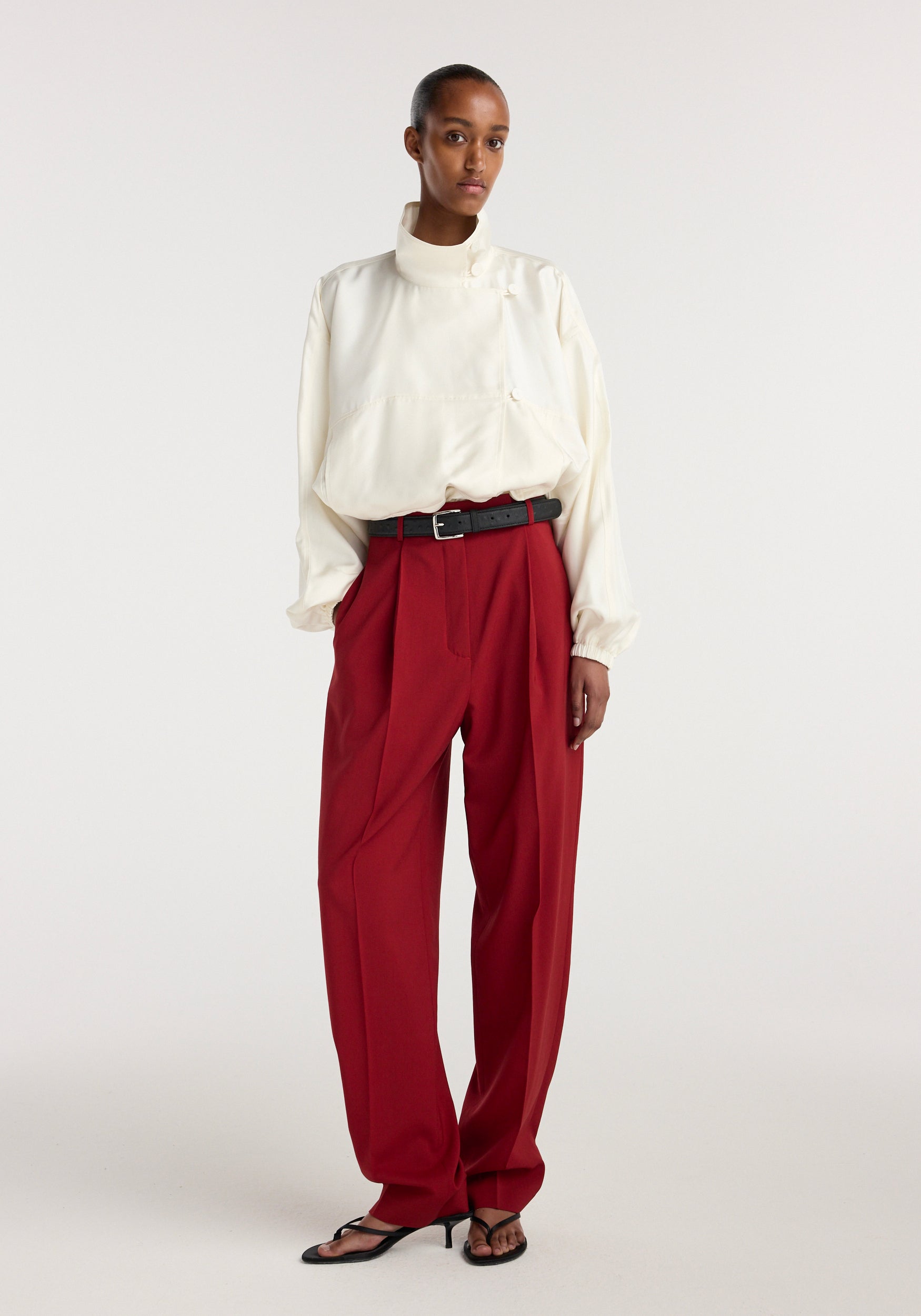 Model wearing Daria - Single pleat tapered trousers | carmine paired with a loose cream blouse and black belt, standing against a plain light background.