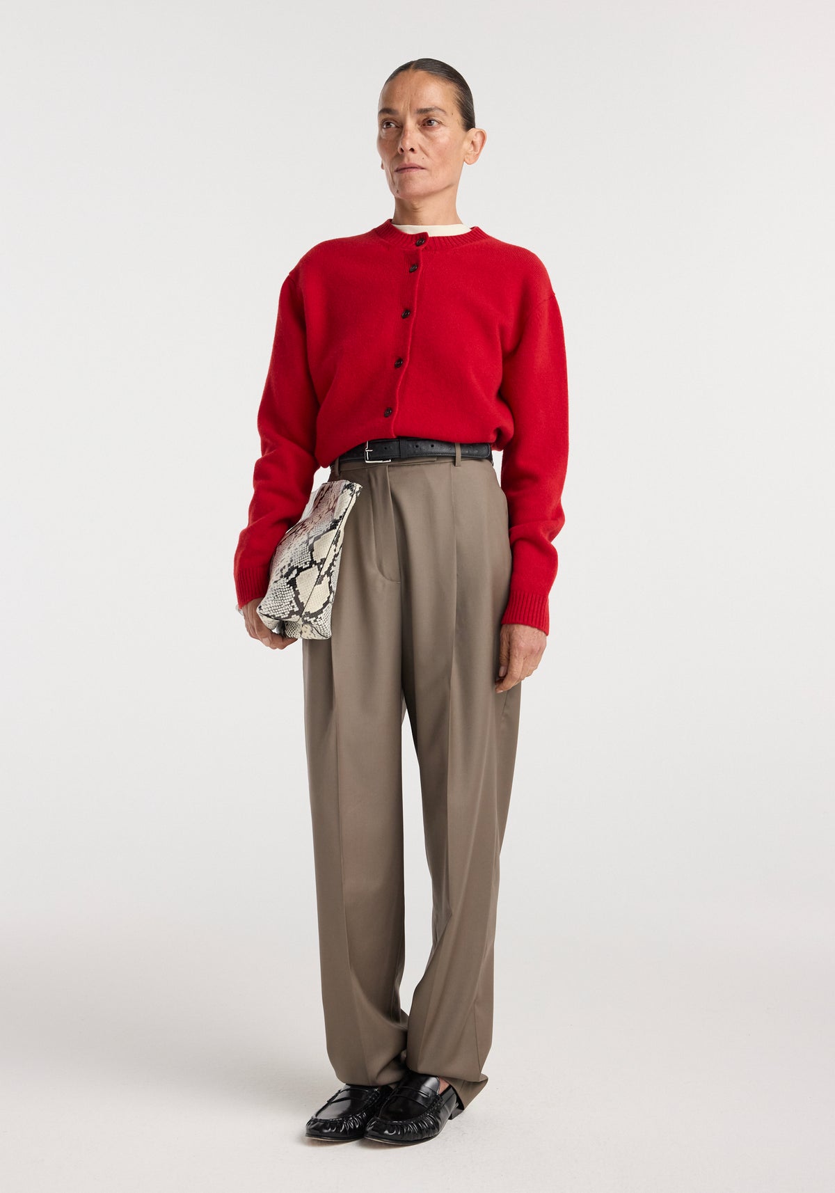 Woman wearing desert brown Daria - Single pleat tapered trousers with a black belt, red button-up sweater, black shoes, and holding a snakeskin-patterned clutch against a white background.