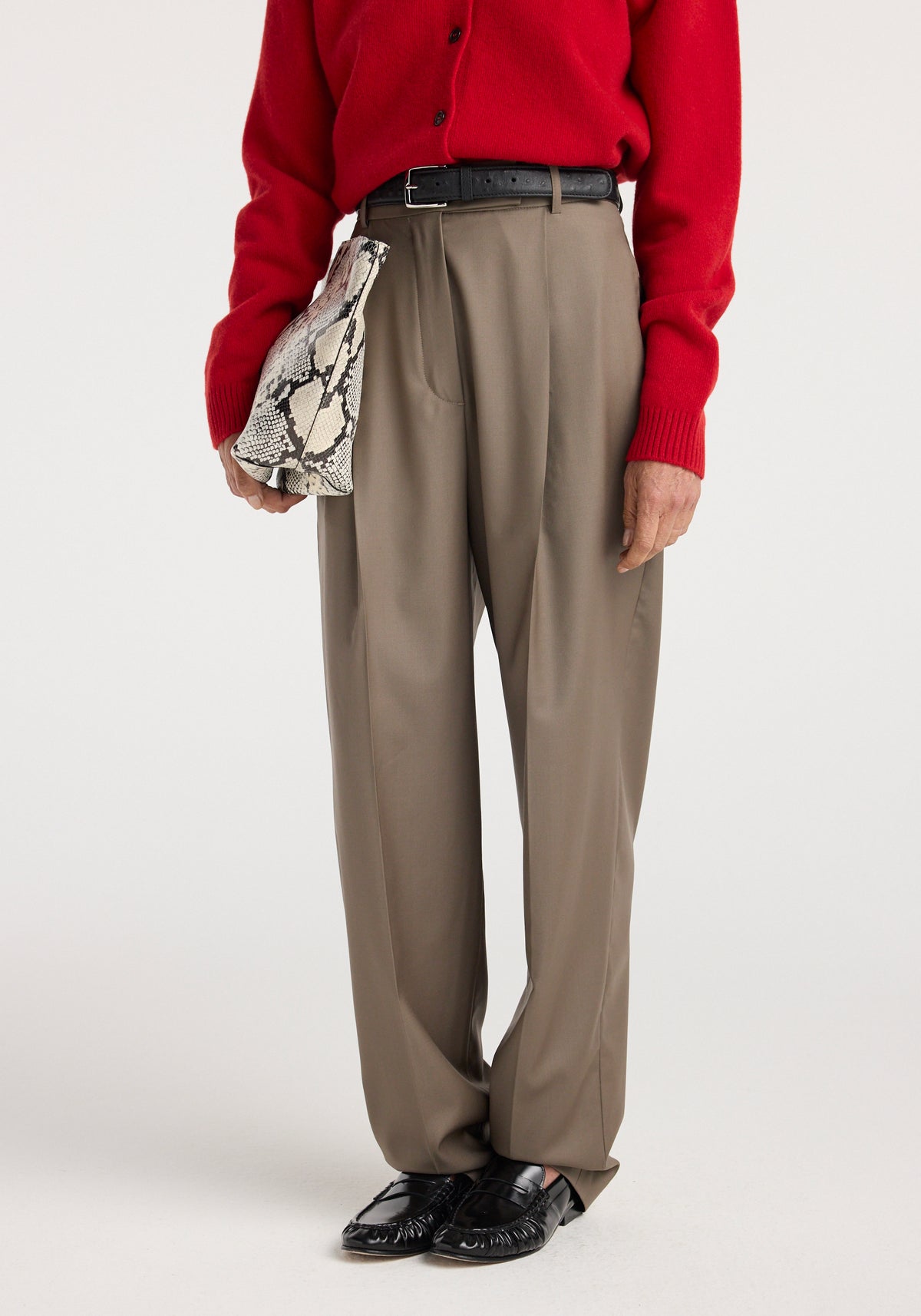Person wearing Daria - Single pleat tapered trousers | desert brown paired with a red sweater, black belt, black shoes, and holding a snakeskin-patterned handbag.