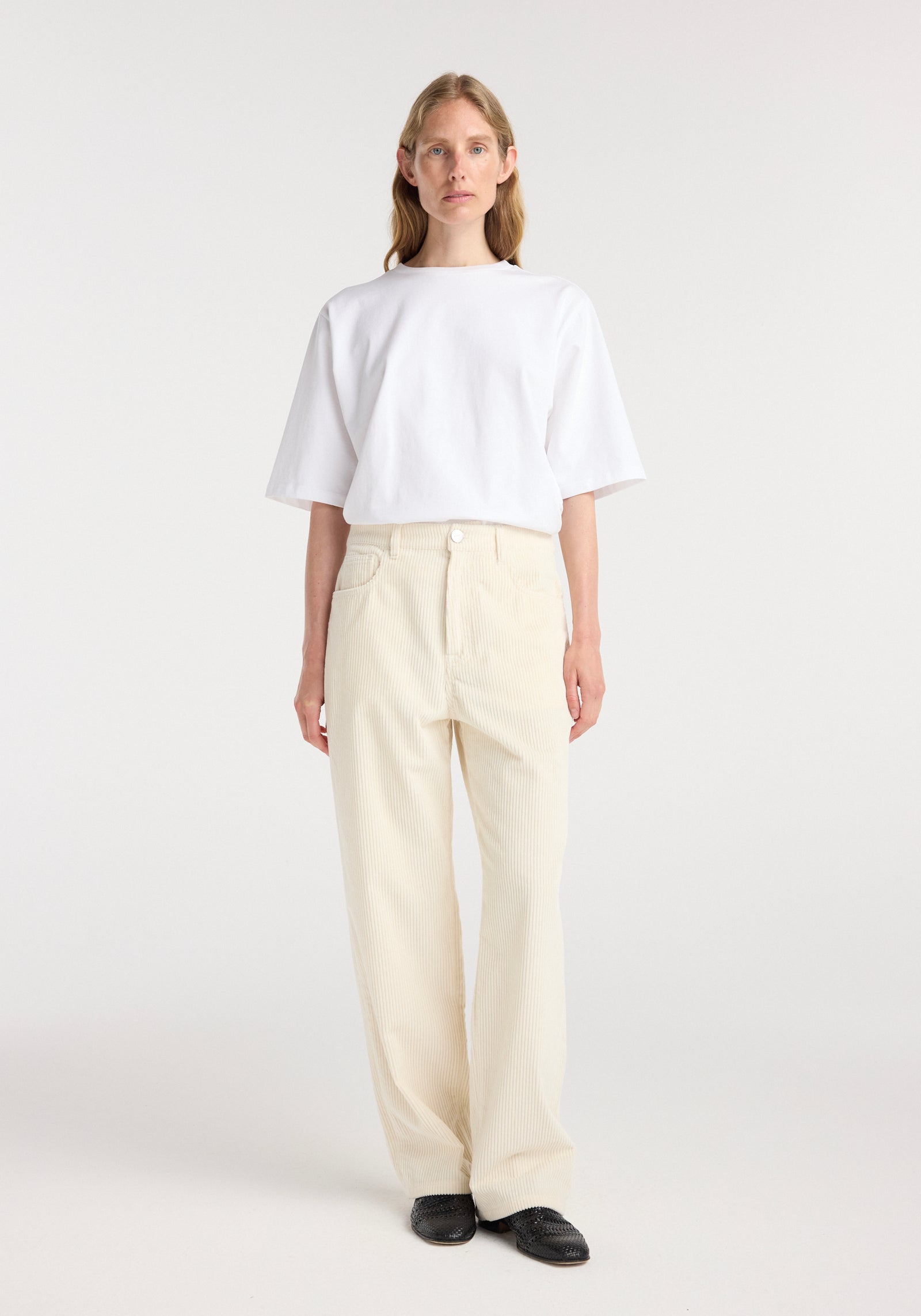 A model wearing Bruun - Signature baggy corduroy | winter white pants paired with a plain white oversized short-sleeve shirt and black shoes, standing against a white background.
