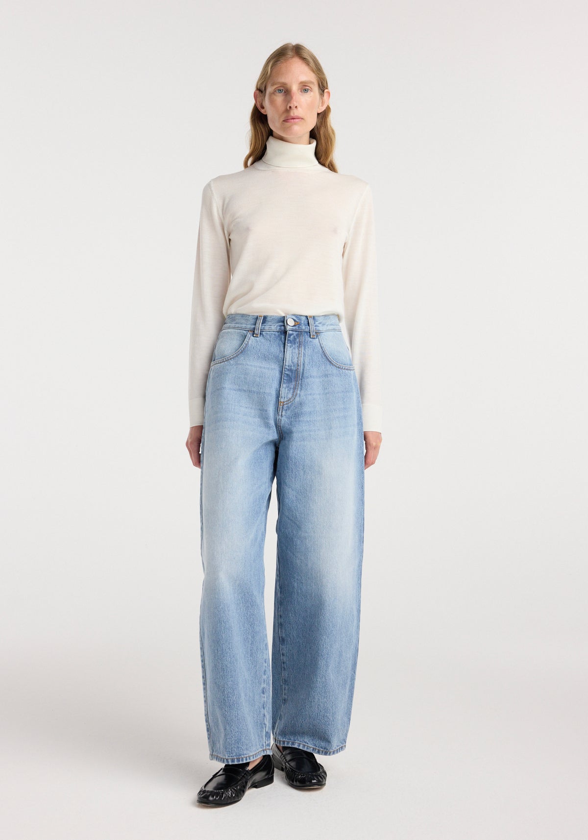 Woman wearing Berber - Barrel leg denim | classic blue jeans paired with a light beige turtleneck sweater and black shoes, standing against a plain background.