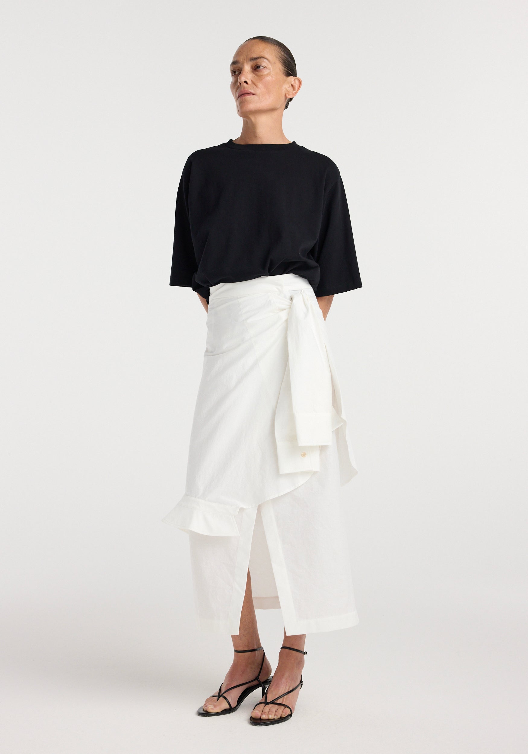 Woman wearing a long optic white shirt-wrap detailed skirt | optic white with a ruffle hem, paired with a loose black short-sleeve top and black strappy heels, standing against a plain background.