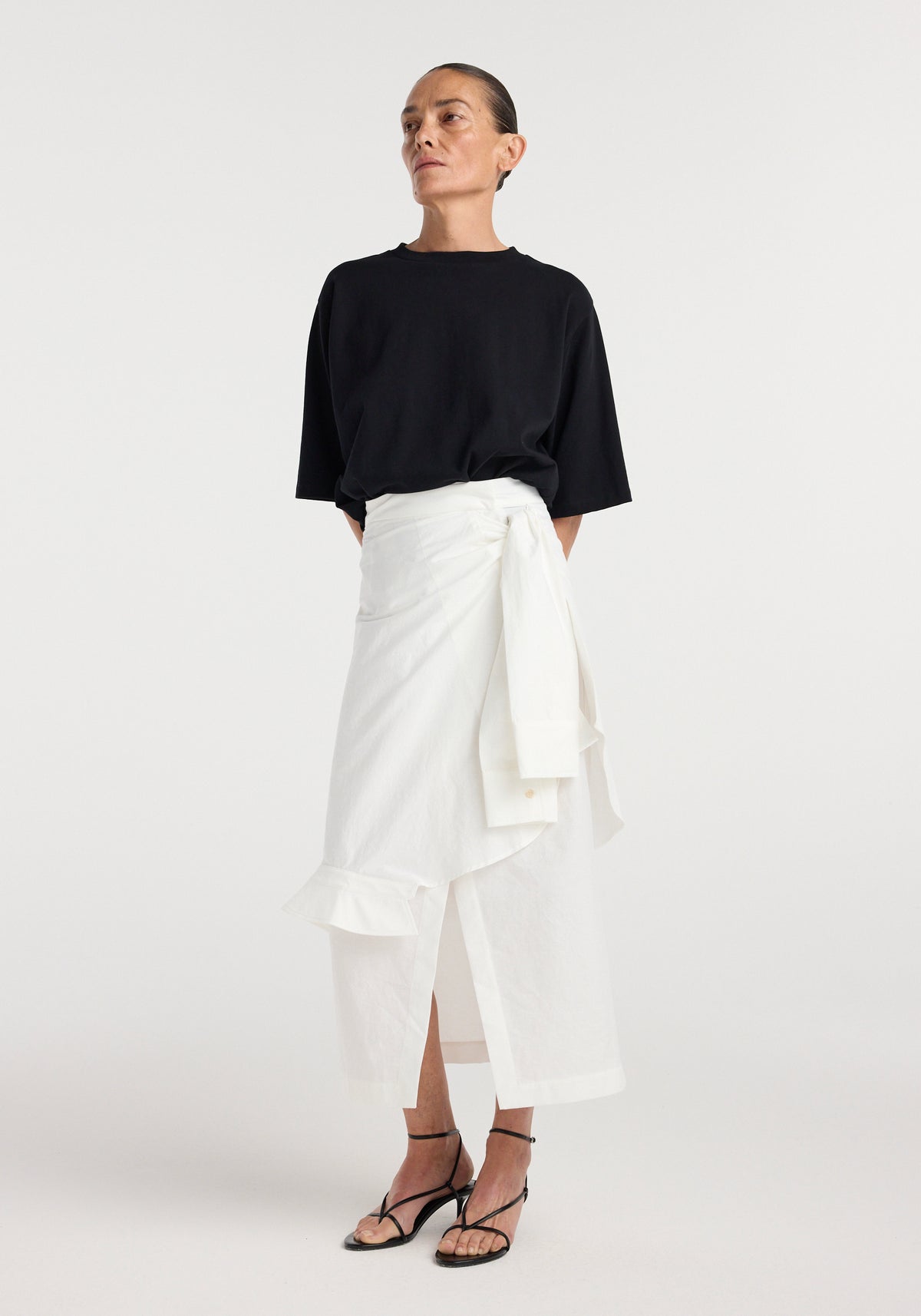 Model wearing the Shirt-wrap detailed skirt | optic white with a black short sleeve top and strappy sandals in a full length studio view
