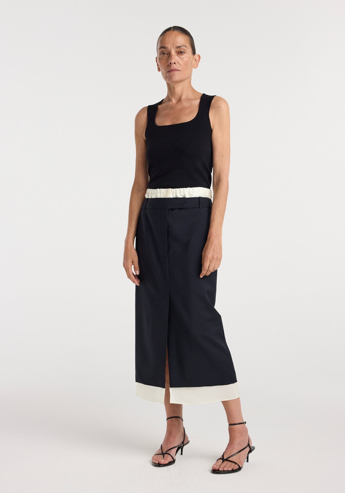 Woman wearing a navy Boxer detailed skirt | navy with a white waistband and hem, paired with a black sleeveless top and black strappy heels, standing against a plain background.