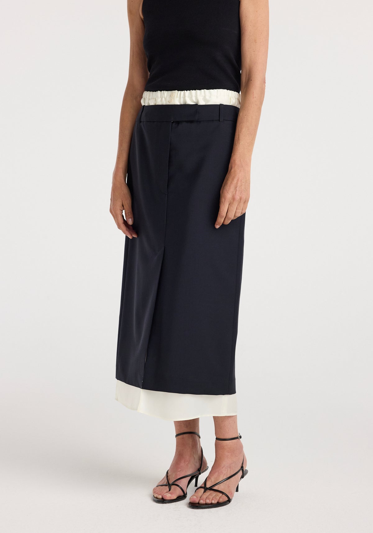 A model wearing a navy Boxer detailed skirt | navy with a white waistband and hem detail, paired with a black sleeveless top and black strappy heeled sandals.