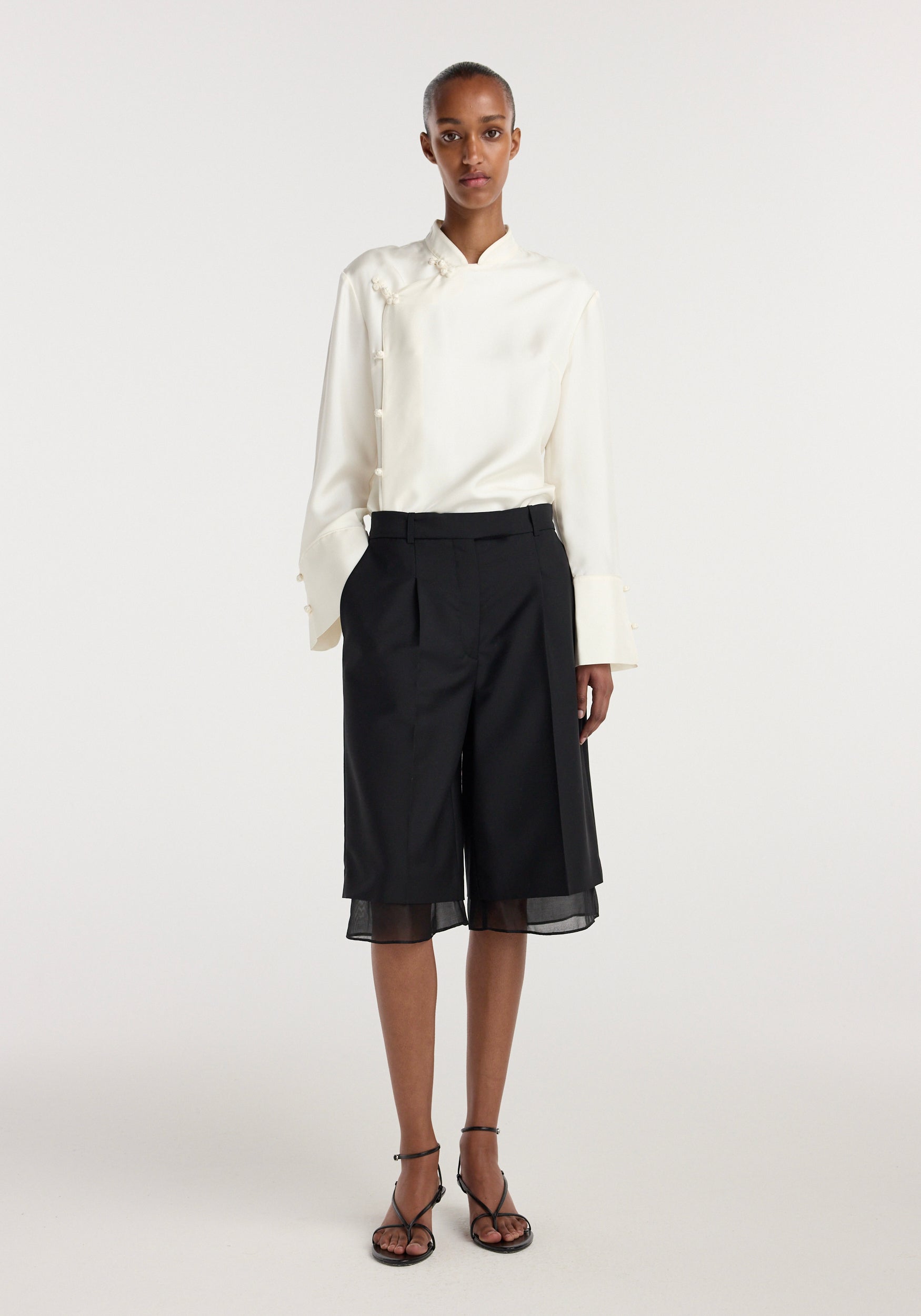 Female model wearing black Organza lined shorts | black paired with a long-sleeve cream blouse and black strappy sandals against a plain light background.