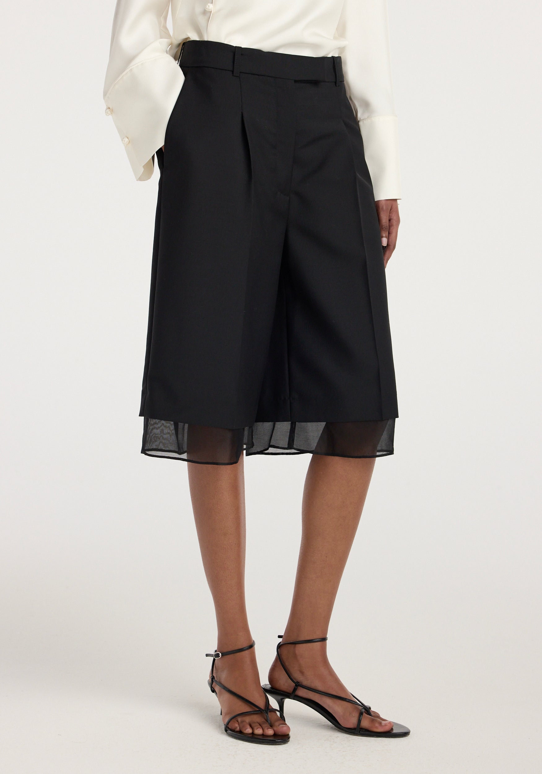 Close-up of a model wearing black Organza lined shorts | black with a white long-sleeve blouse and black strappy heels, highlighting the sheer organza layer beneath the shorts.