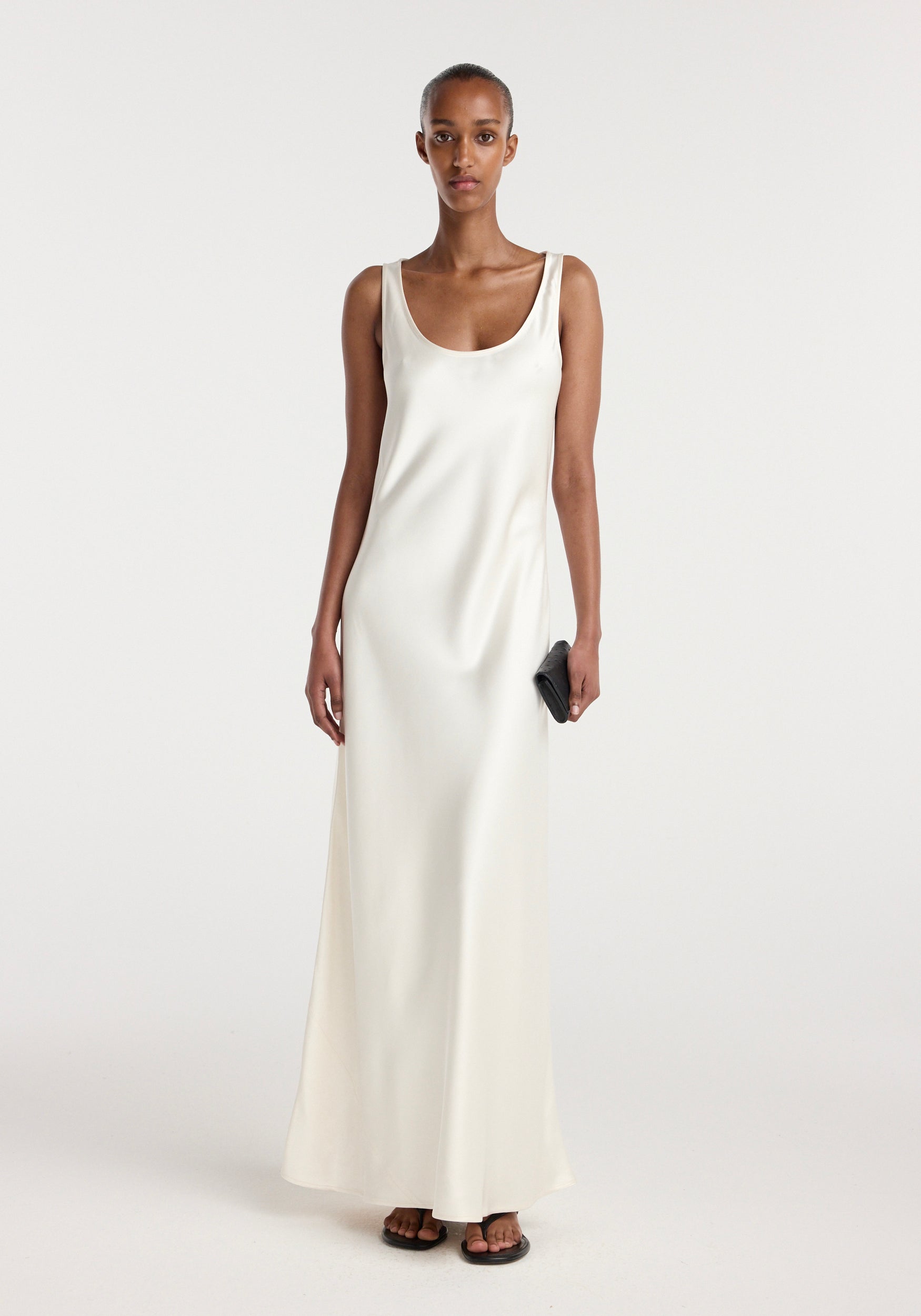 A woman wearing a sleeveless scoop neck satin dress in ivory, standing and holding a small black clutch, paired with black open-toe sandals in a minimalist setting.