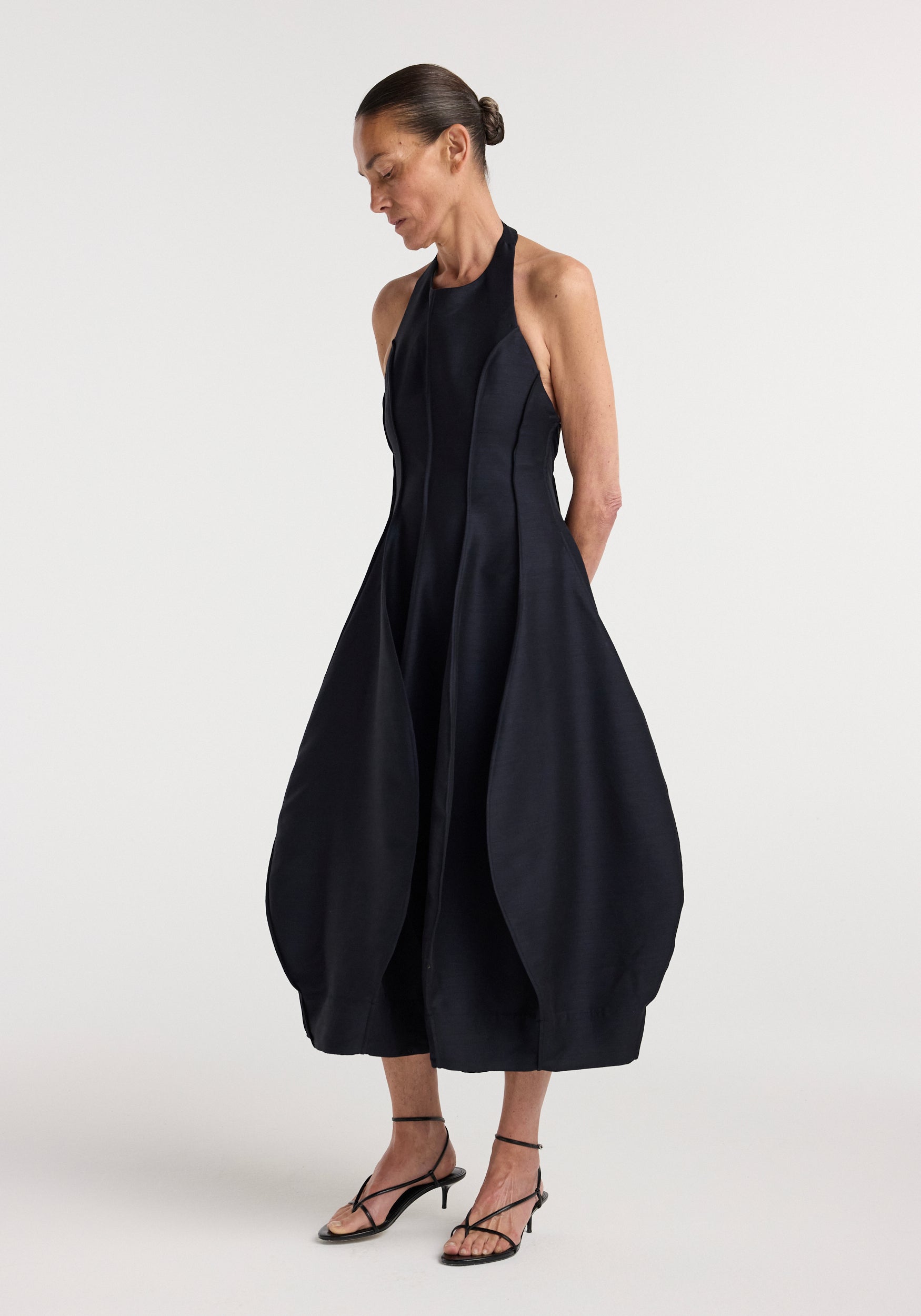 Woman wearing a sleeveless, fitted black open back tulip dress | night with a halter neckline and voluminous tulip-shaped skirt, paired with black strappy heels.