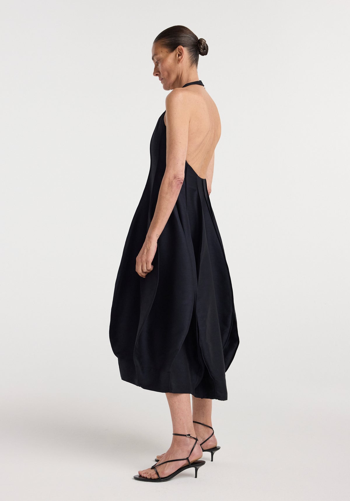 Woman wearing a black open back tulip dress | night with a halter neckline and asymmetrical tulip-shaped skirt, paired with black strappy kitten heels against a plain background.
