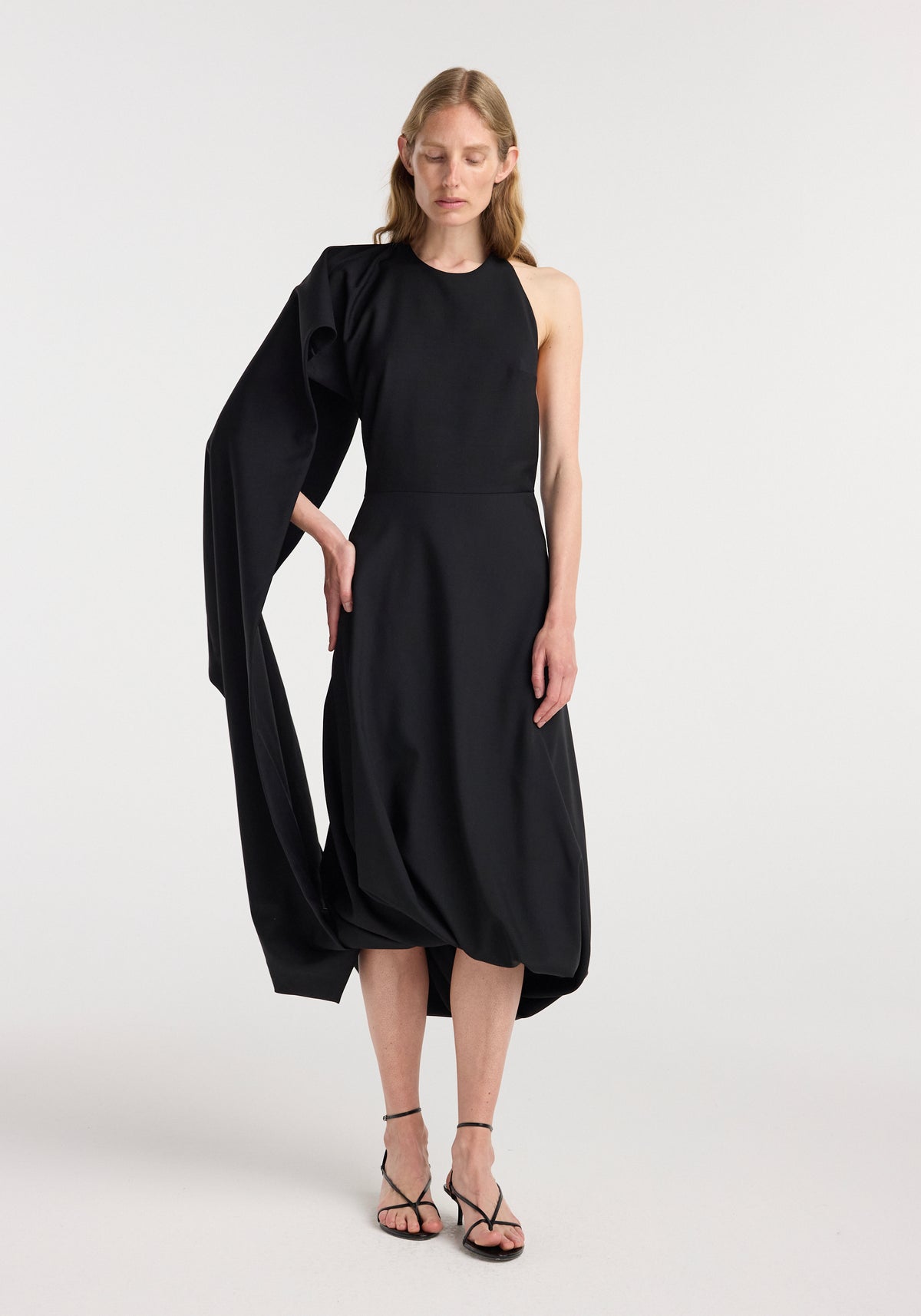 Woman wearing a black twisted cape dress | black with a draped, asymmetrical cape detail on one shoulder and a midi-length skirt, paired with black strappy sandals.