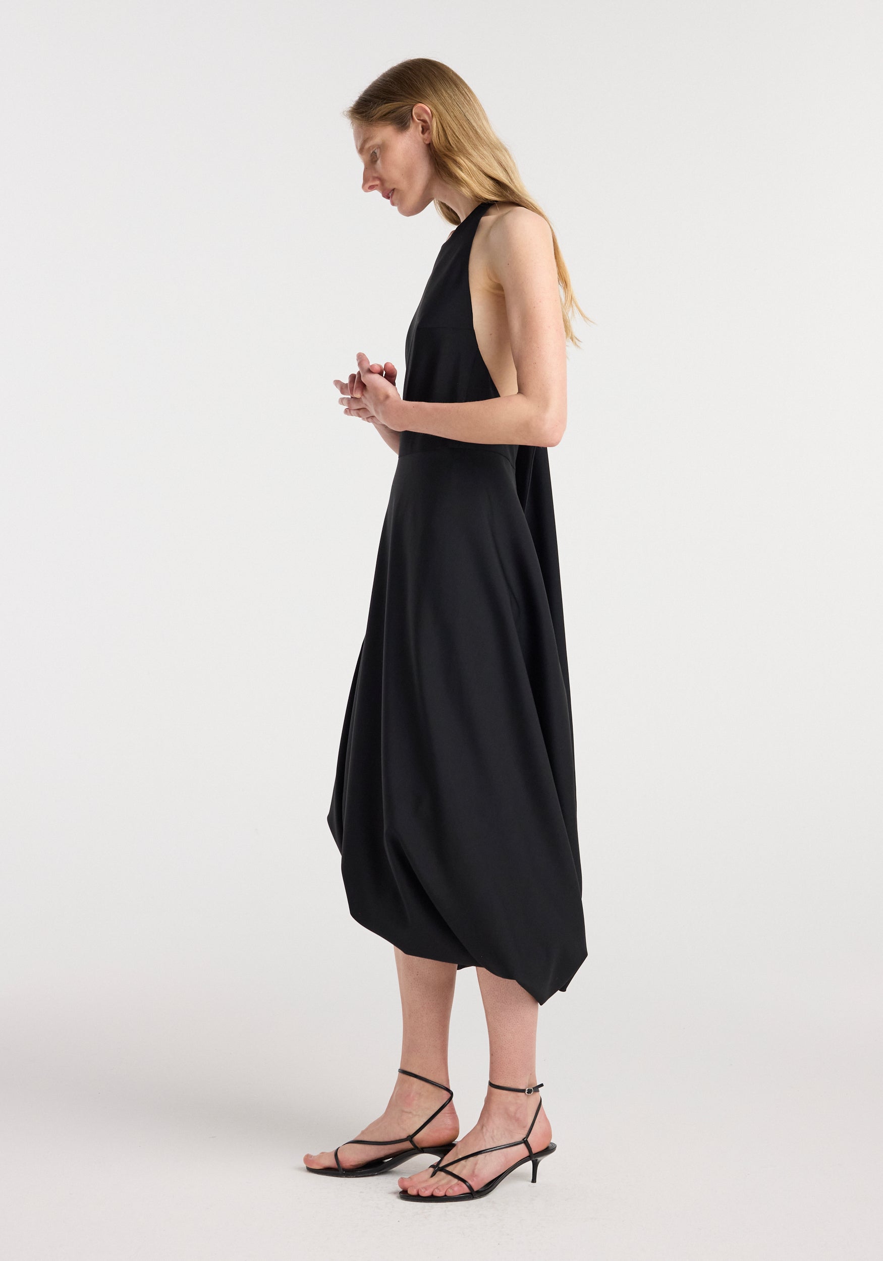 Side profile of a woman wearing a loose, sleeveless black twisted cape dress | black with a high neckline, paired with black strappy low-heeled sandals against a plain white background.