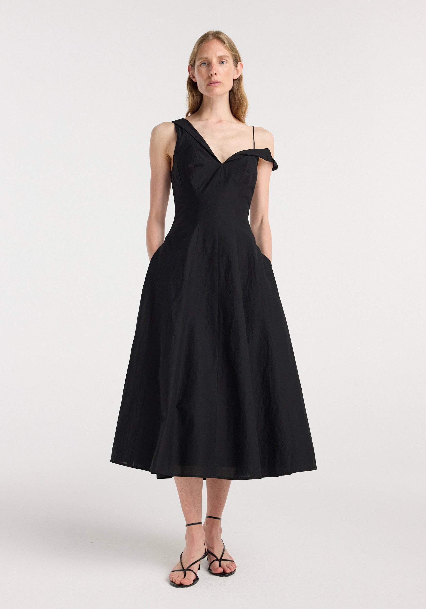 Woman wearing a black twisted shoulder dress | black with one off-shoulder strap and a fitted waist, standing against a white background with hands in pockets.