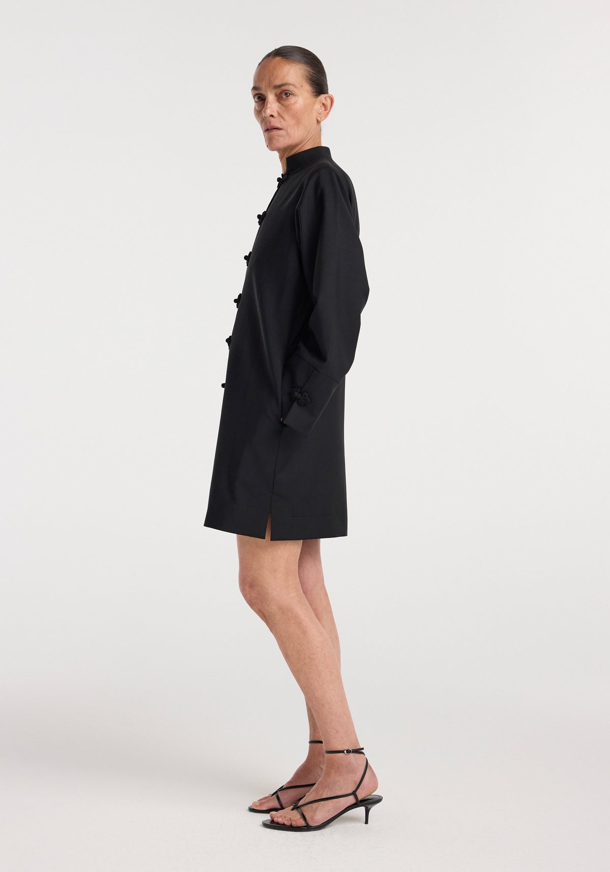 Woman wearing a black Pankou closure dress | black with long sleeves, a high collar, and a straight cut, paired with black strappy heeled sandals.