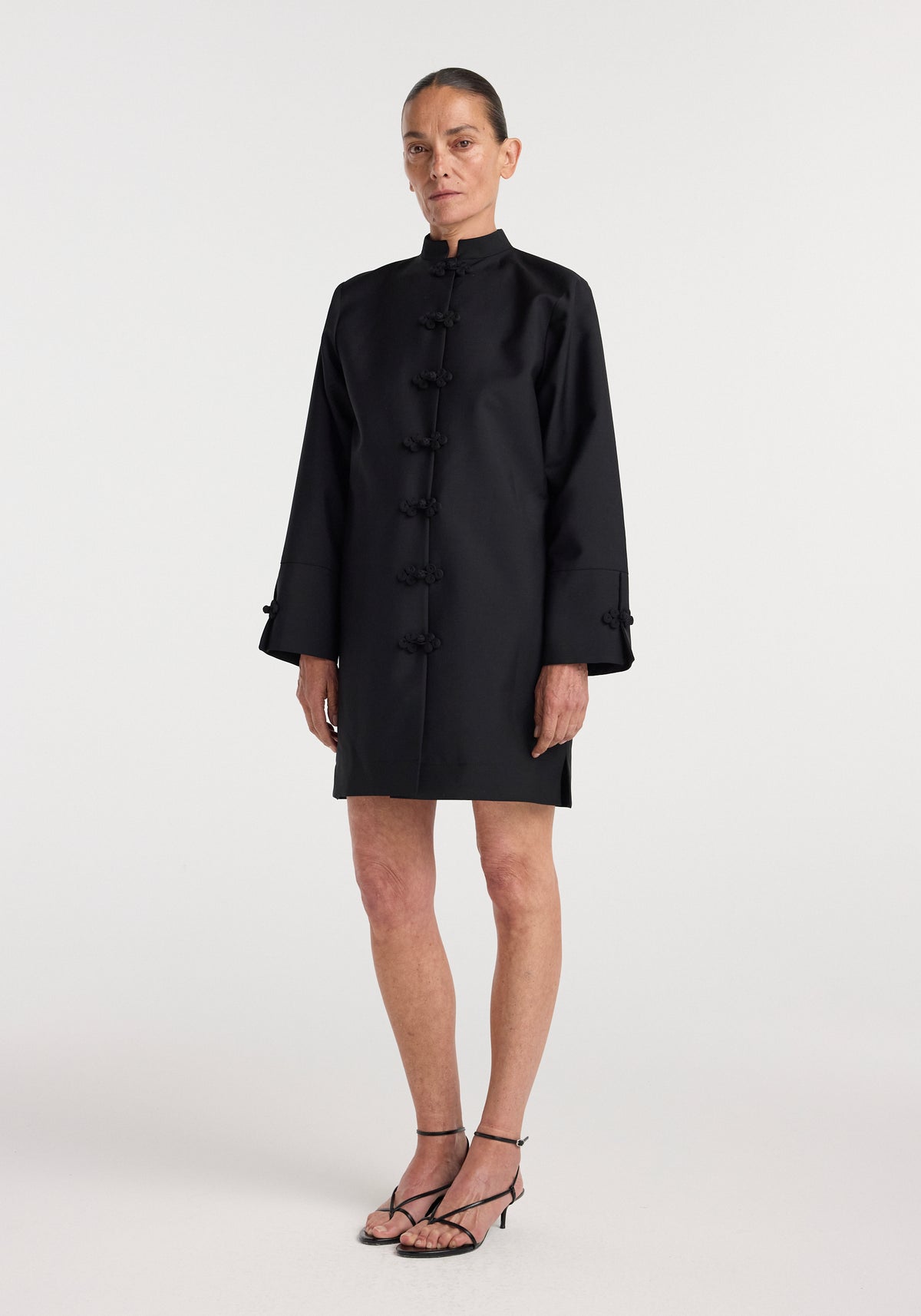 Woman wearing a black Pankou closure dress | black with long sleeves and traditional knot buttons down the front, paired with black strappy sandals, standing against a white background.