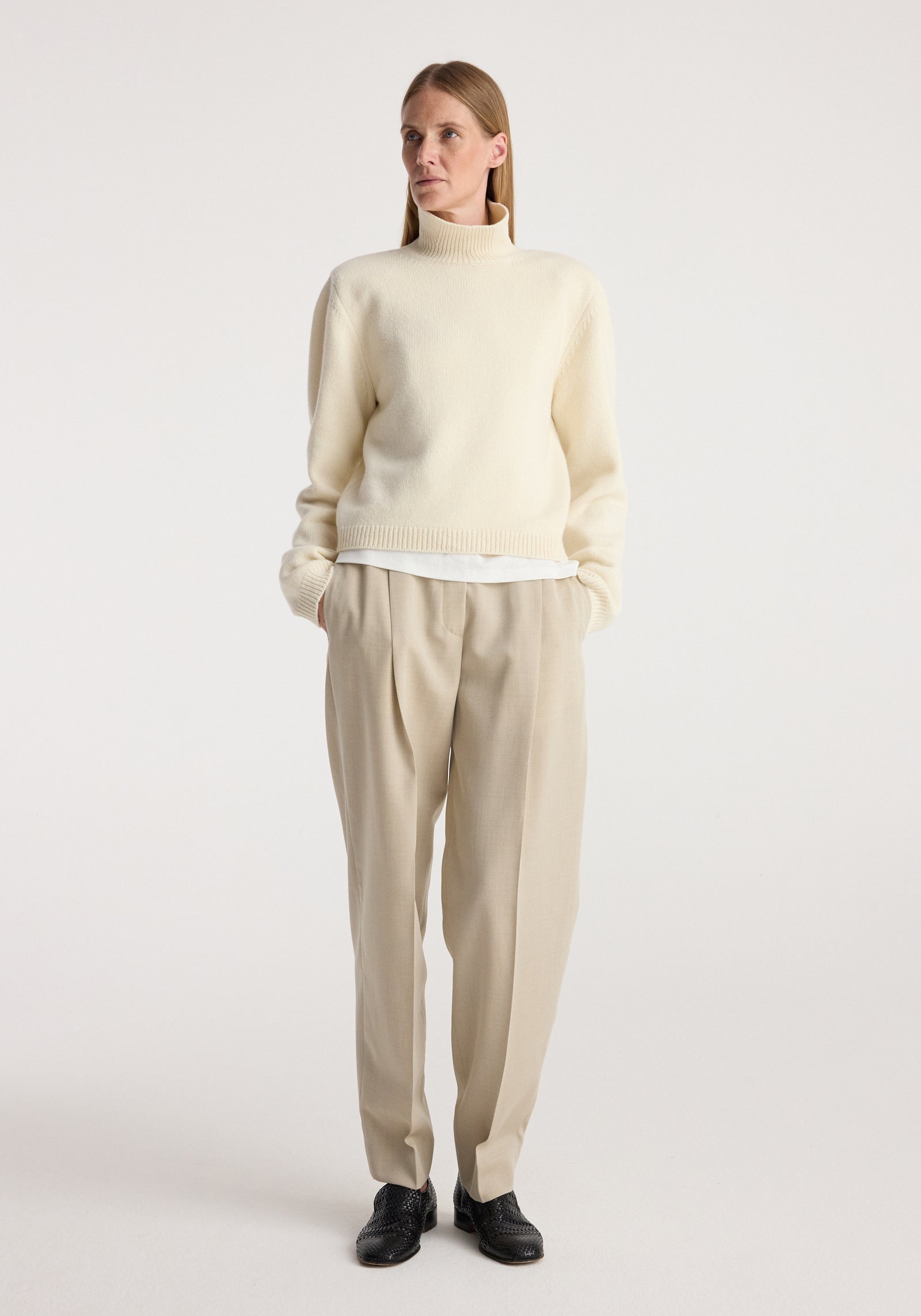 Woman wearing a cream turtleneck sweater with statement shoulders and cropped length, styled with beige pleated trousers and black shoes, standing against a plain background, Statement shoulder cropped turtleneck | cream.
