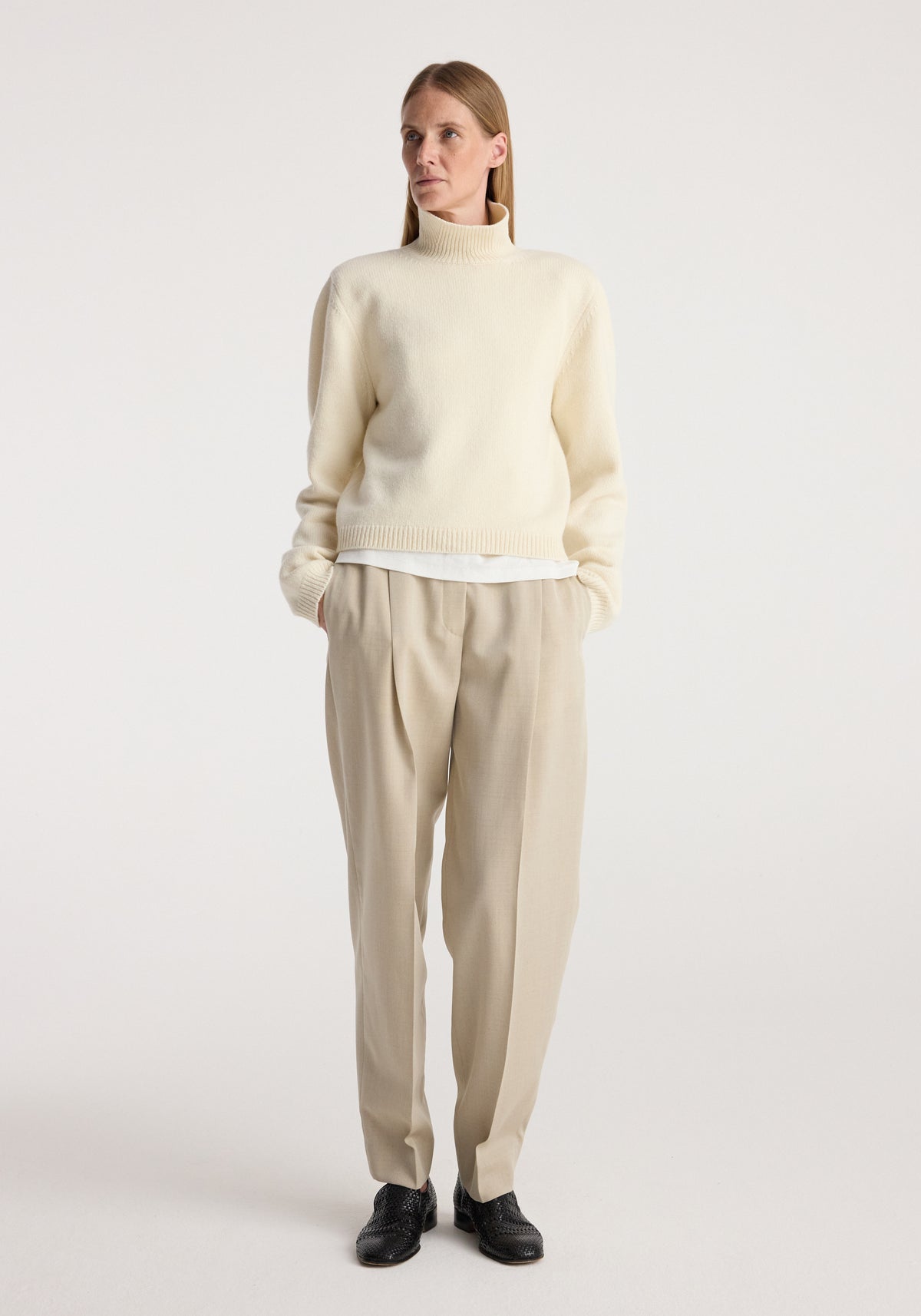 Woman wearing a cream turtleneck sweater with statement shoulders and cropped length, styled with beige pleated trousers and black shoes, standing against a plain background, Statement shoulder cropped turtleneck | cream.