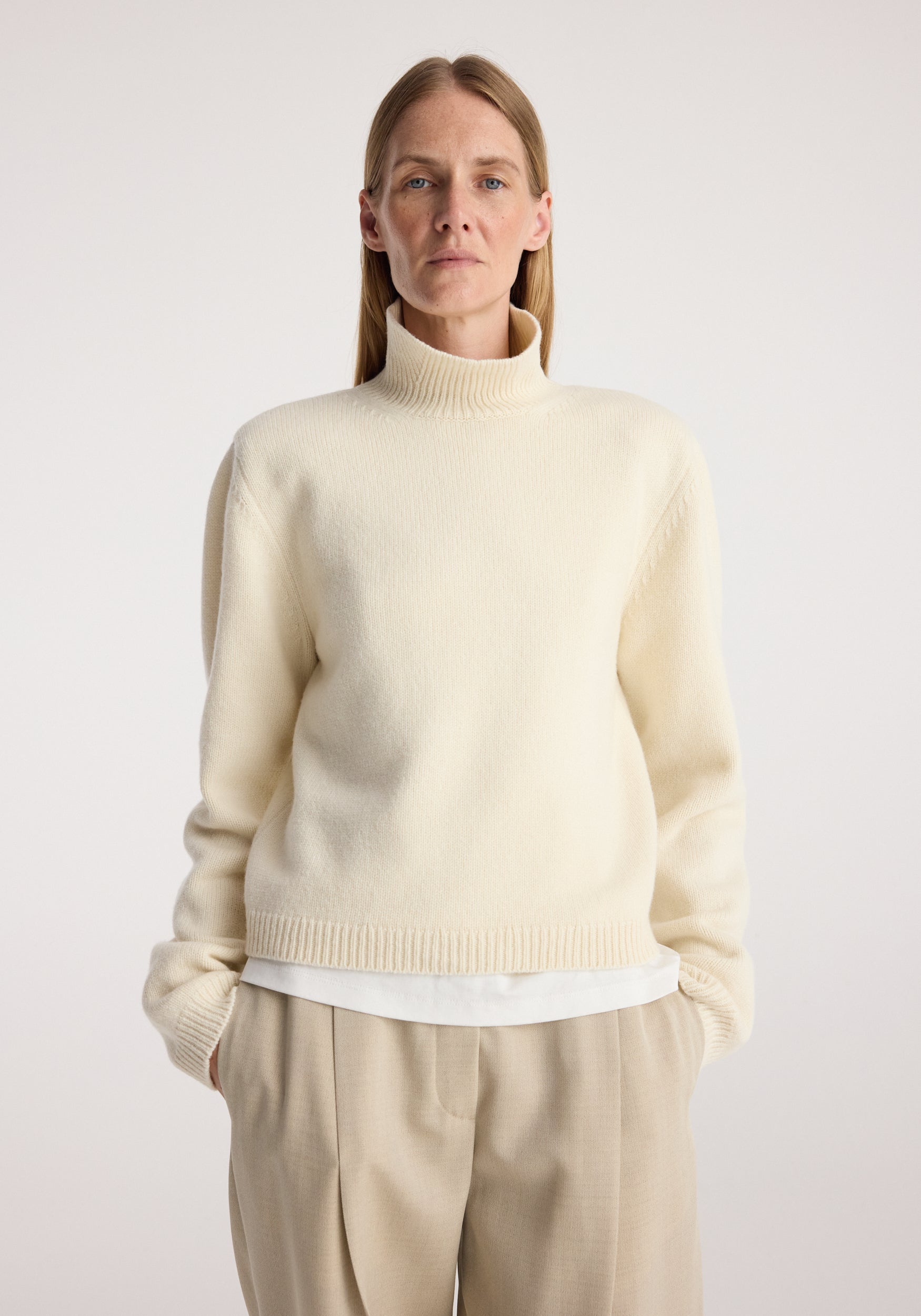 Woman wearing a cream Statement shoulder cropped turtleneck | cream with long sleeves and a fitted high neck, styled with beige pants against a plain background.