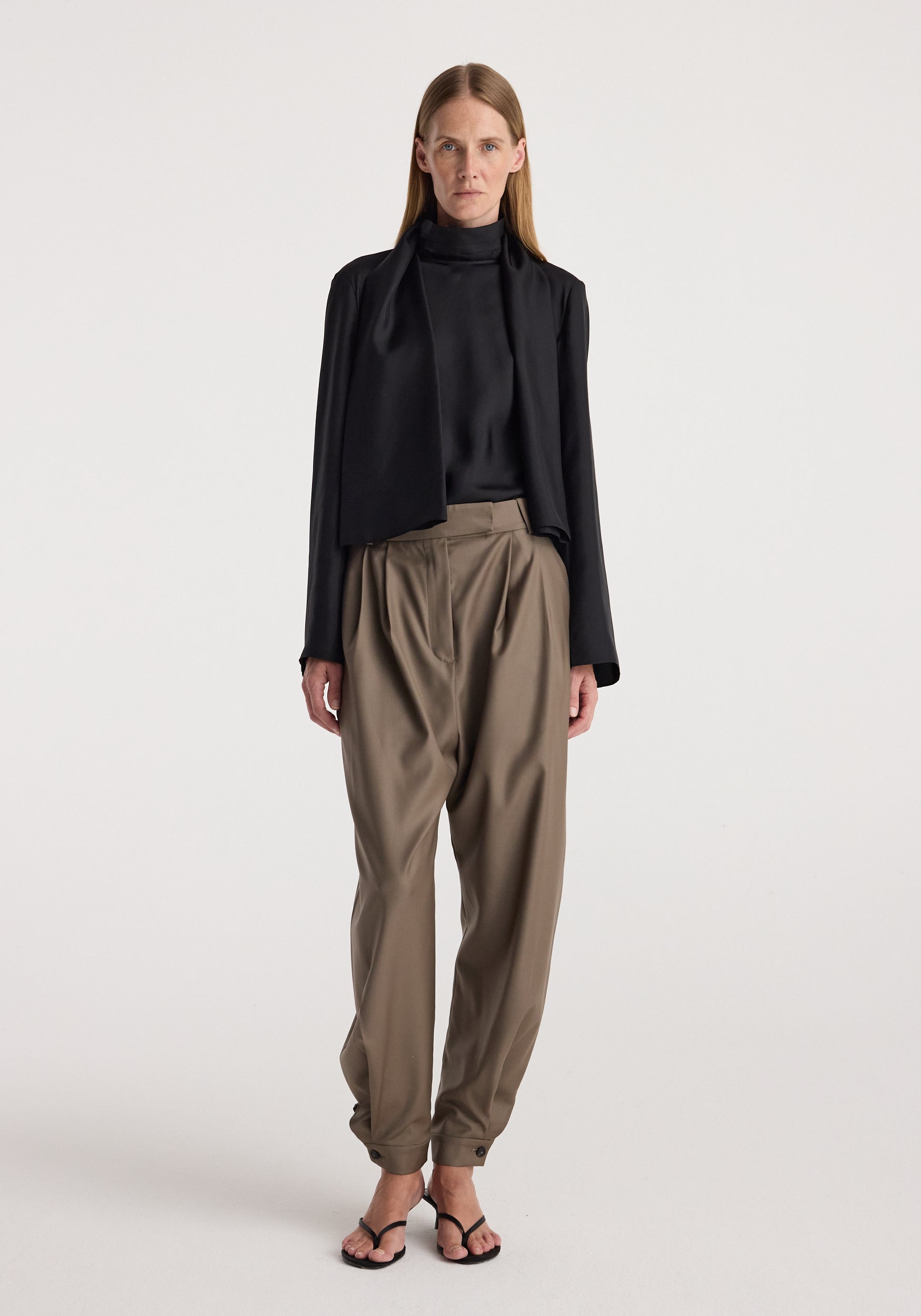 Model wearing tailored cuff trousers | desert brown with a high-waisted fit and pleated front, paired with a black blouse and black cropped jacket against a plain background.