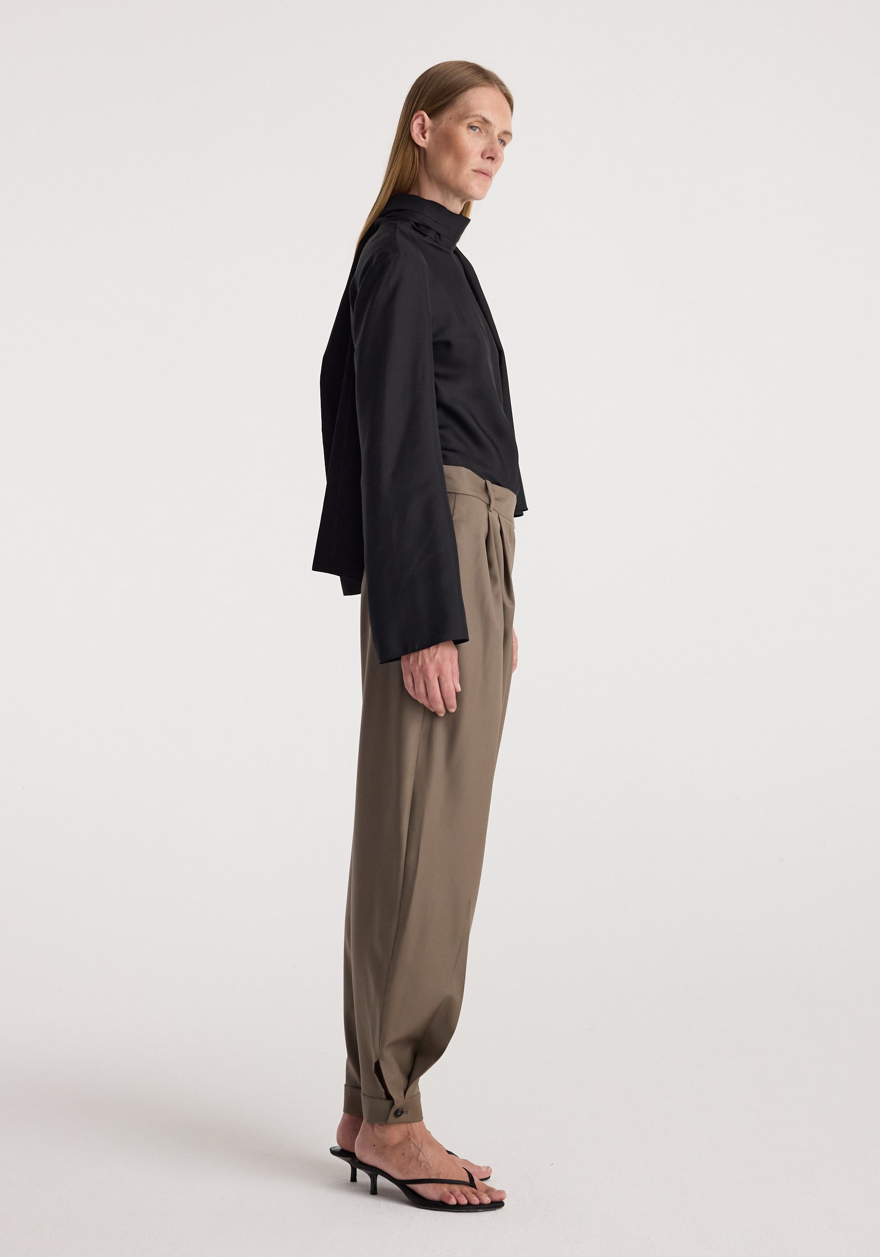 Woman wearing tailored cuff trousers | desert brown styled with a black long-sleeve top and black heeled sandals, standing in profile against a plain background.